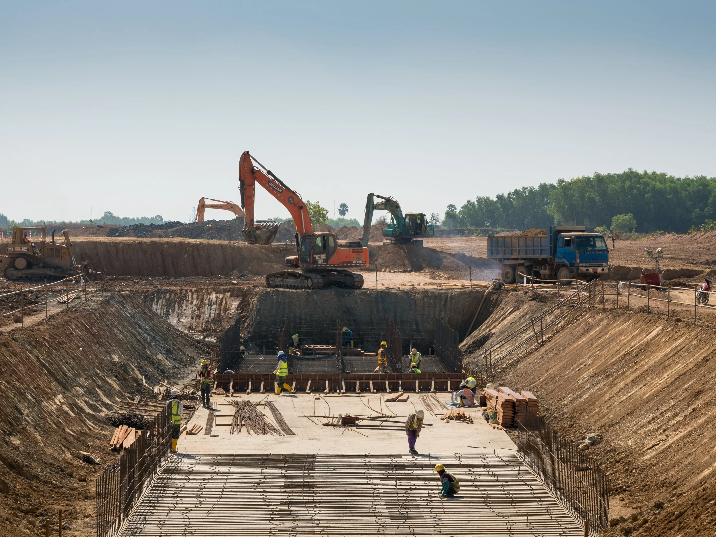 Construction site with workers installing reinforced steel bars in a deep excavation, heavy machinery including excavators and trucks, and a clear sky in the background.