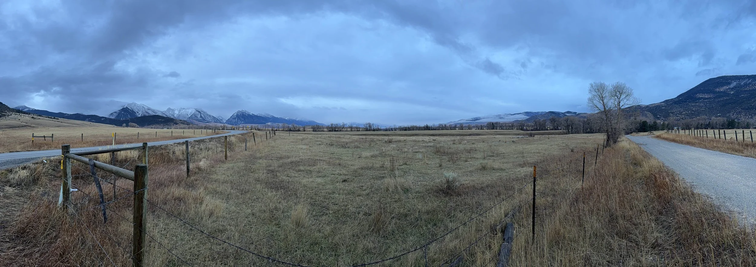 Open Montana prairie with dry grass, fence, and snow-capped mountains under cloudy sky – Cradle of Life LLC