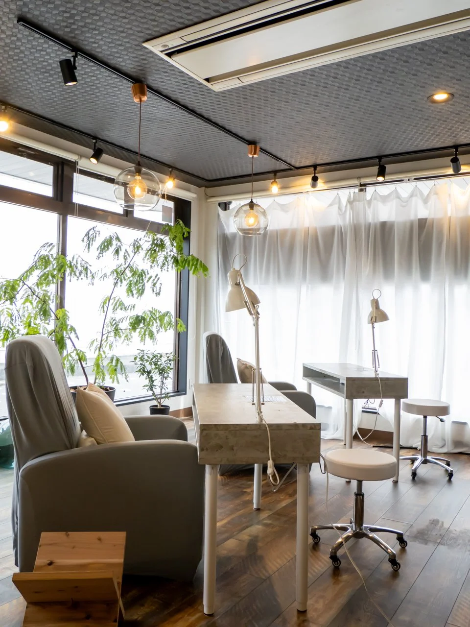 明るい自然光が差し込む、モダンなワークスペース。観葉植物とデスク、チェア、スタンドライトが配置されている。天井に照明と空調設備がある。