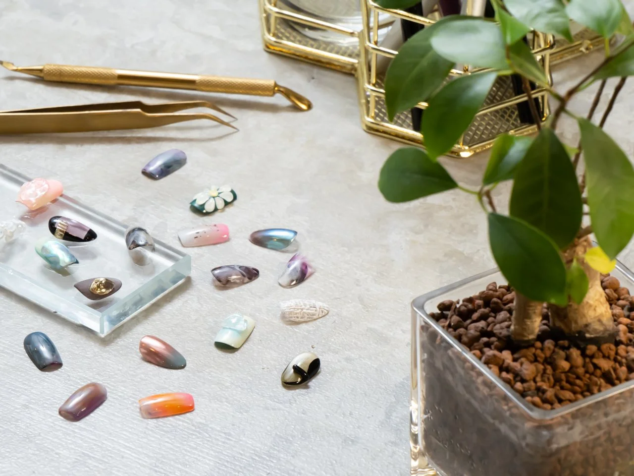 ガラス製のネイルパーツがテーブル上に散らばっている。右側には緑の葉と木が入った透明な花瓶が置かれている。左側には金色のピンセットと細工棒が置かれている。