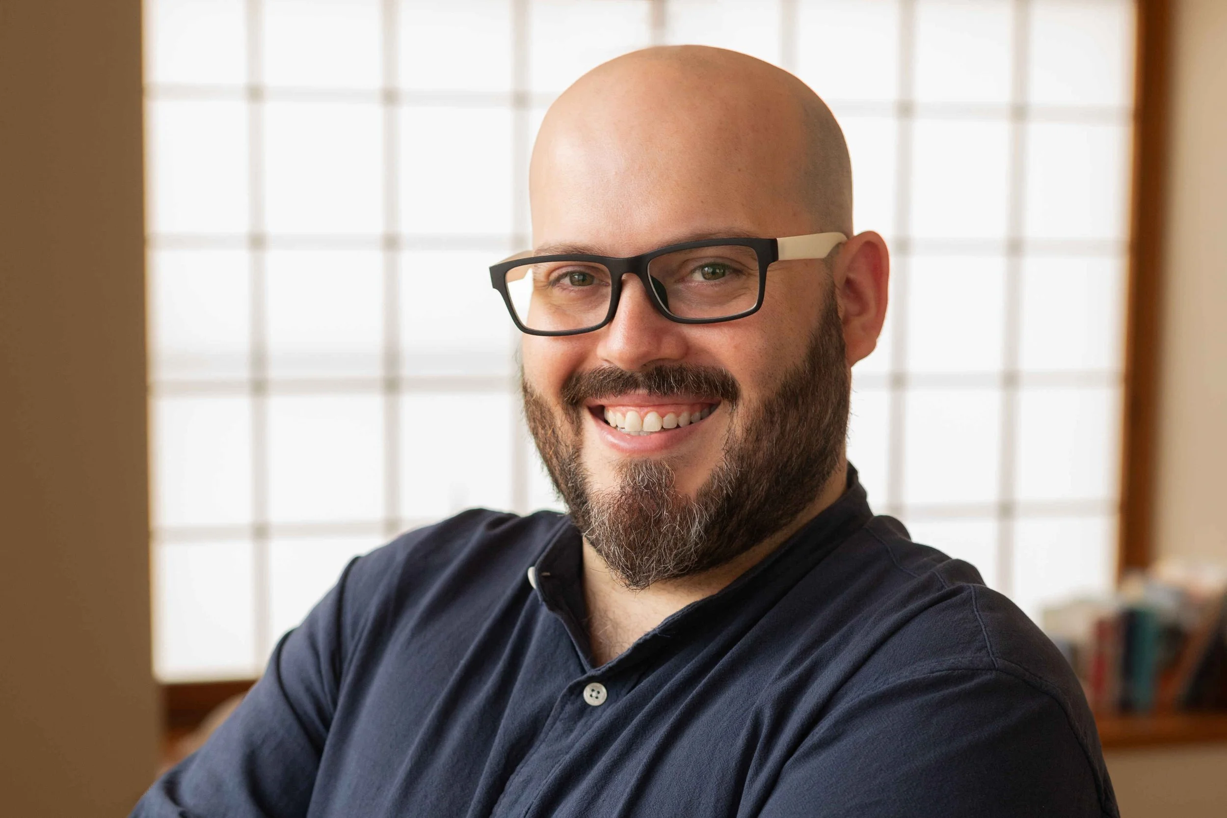A smiling man with a beard and glasses looking at a camera, with a window with grid pattern behind him and blurred bookshelves to the side.