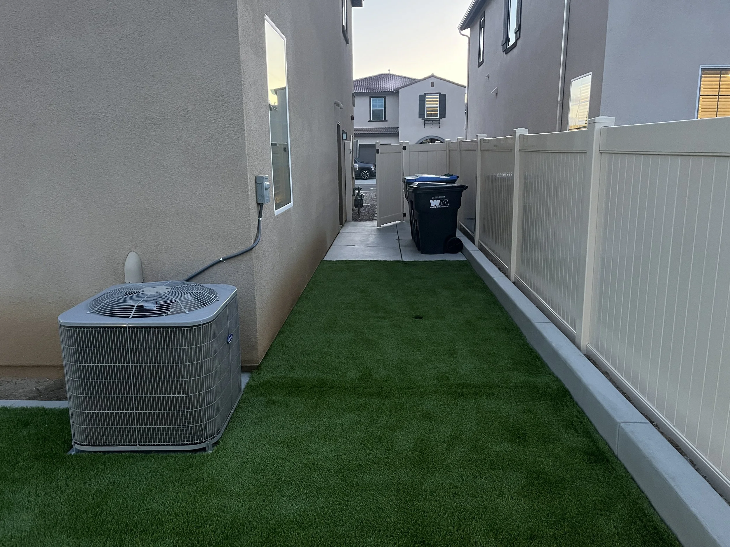 Side yard with artificial grass, an air conditioning unit, two black trash bins, and a white vinyl fence with neighboring houses in the background.