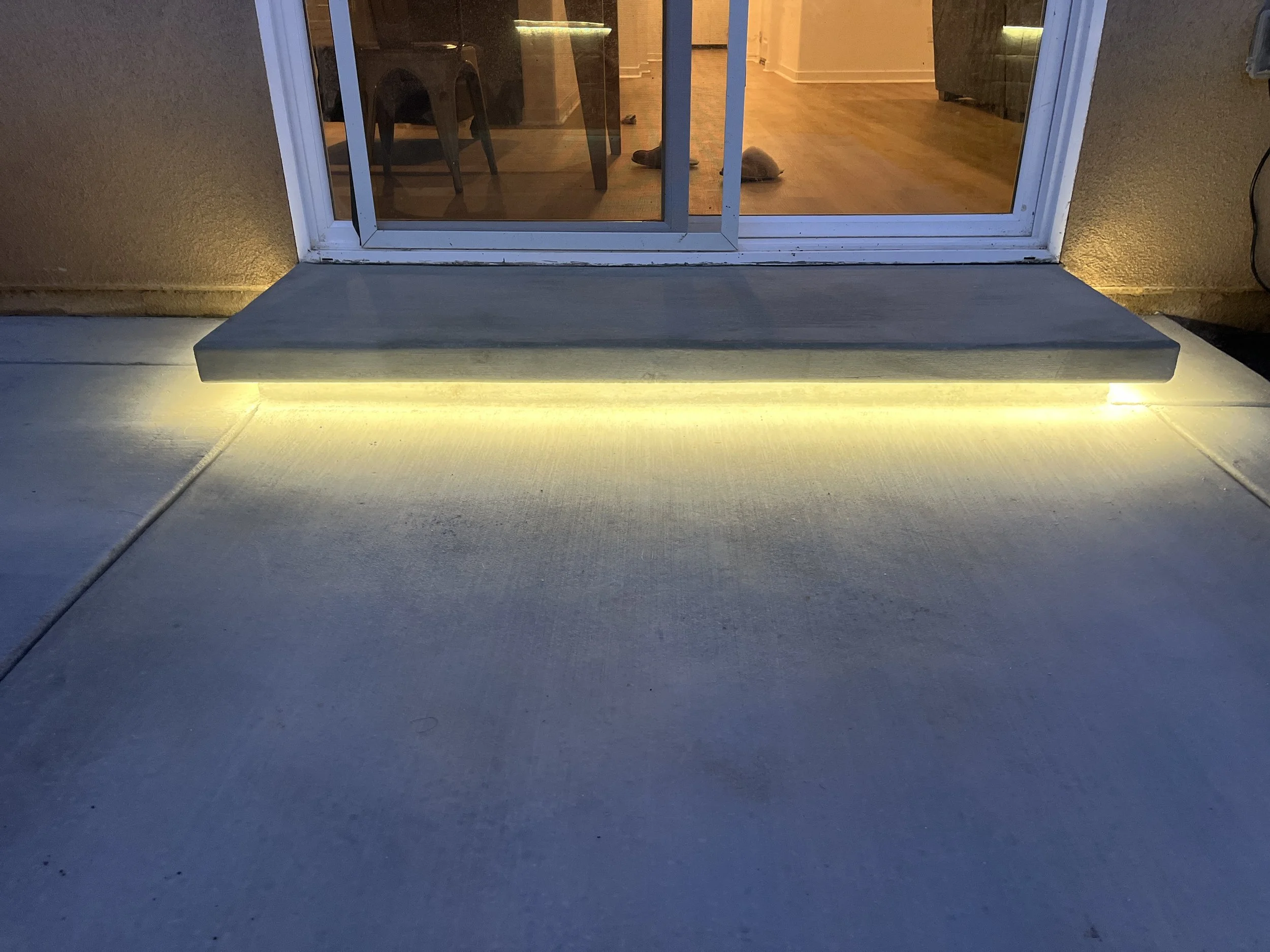 The photo shows a door with a small staircase leading to it. There are yellow LED lights under the stair, illuminating the concrete ground outside the door during nighttime. The interior of the house is visible through the glass door, revealing furni