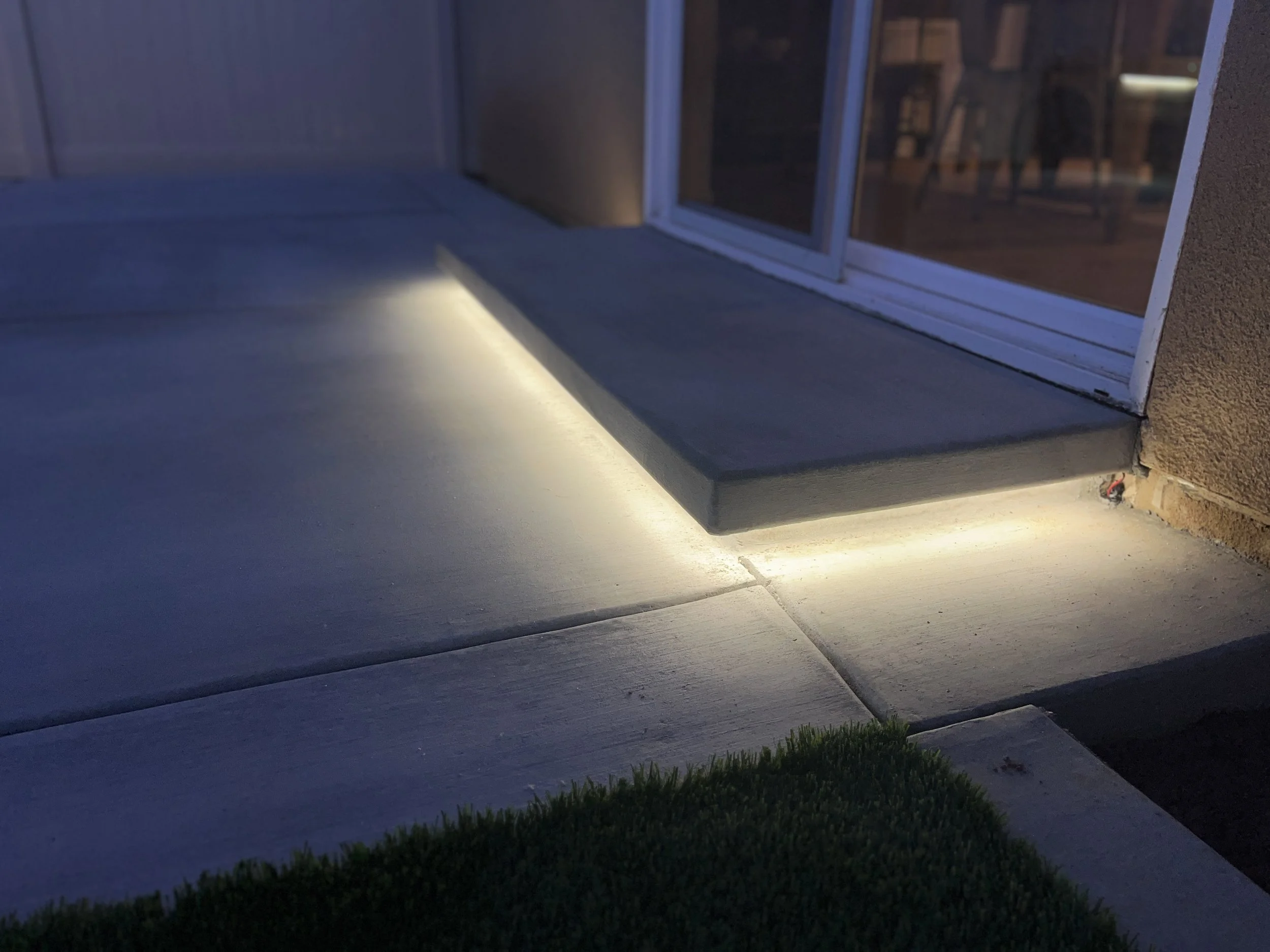 Exterior view of a house entrance at dusk, showing a concrete walkway with installed LED lighting along the edge near a glass door.