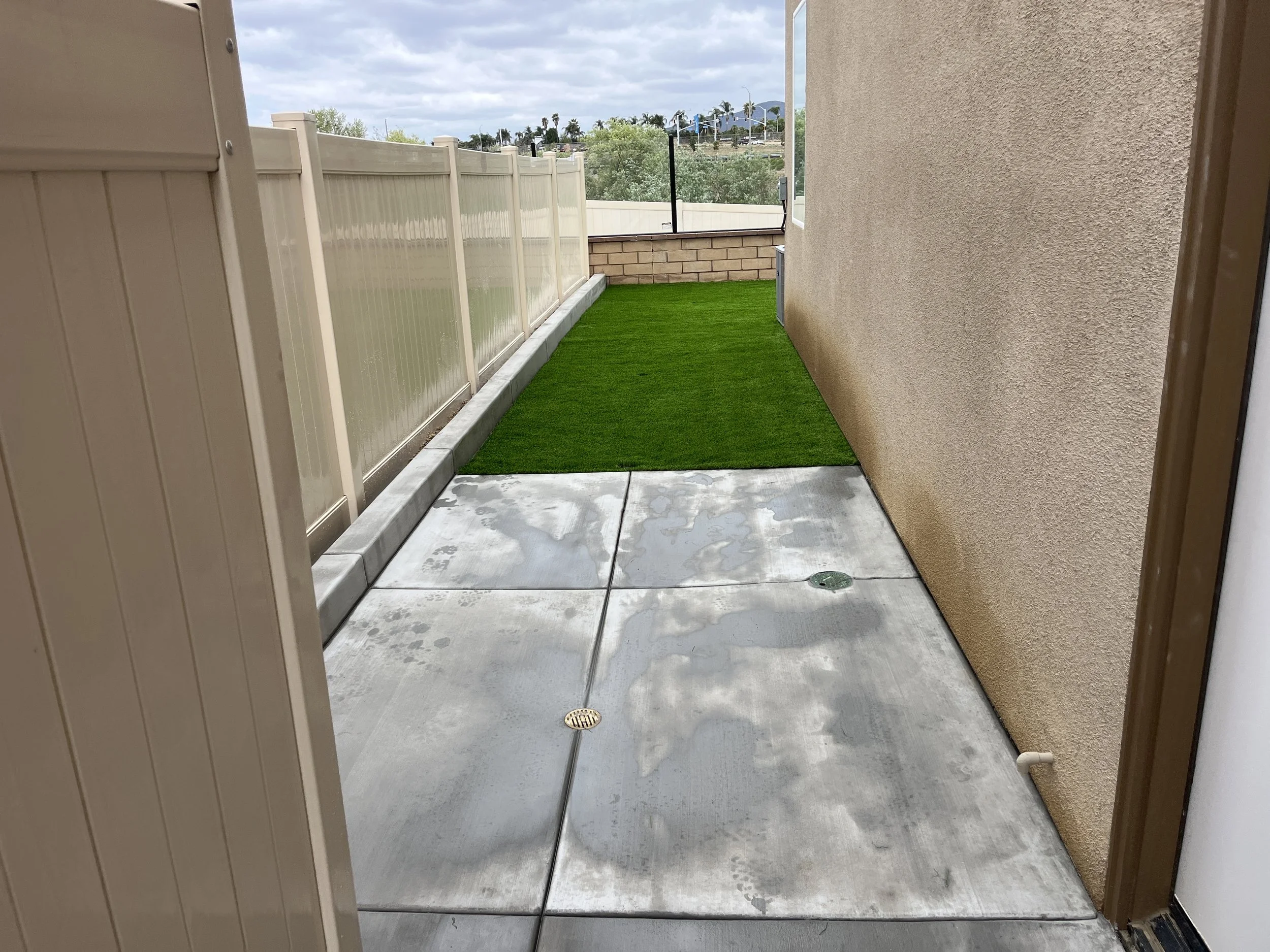 Small outdoor patio with concrete flooring, a patch of artificial grass, beige wall on the right, cream-colored fence on the left, and a view of trees, hills, and cloudy sky in the background.