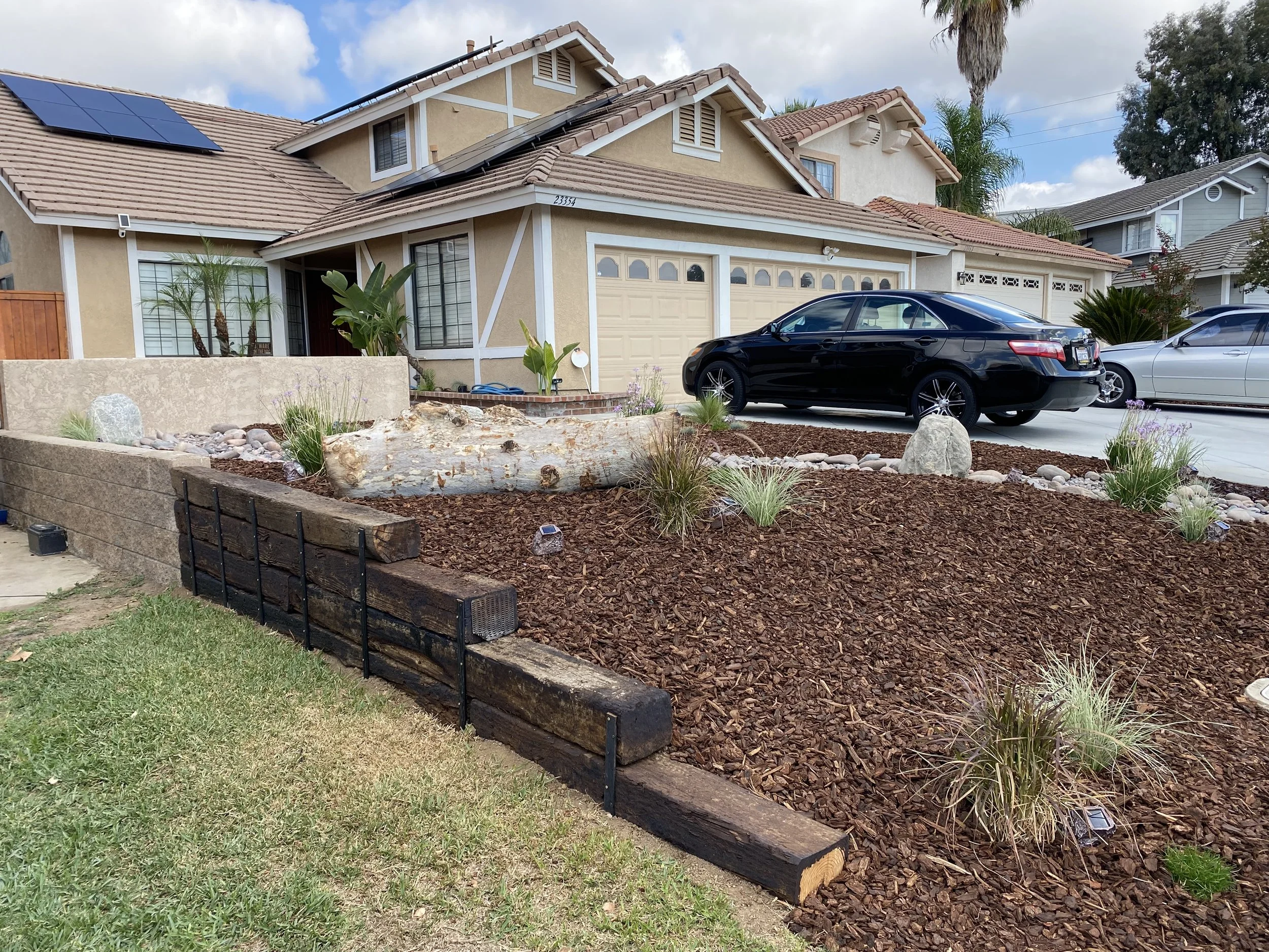 A residential driveway with a black car parked in front of a house, landscaped with a mulch bed, rocks, and small plants, with neighboring houses visible in the background.