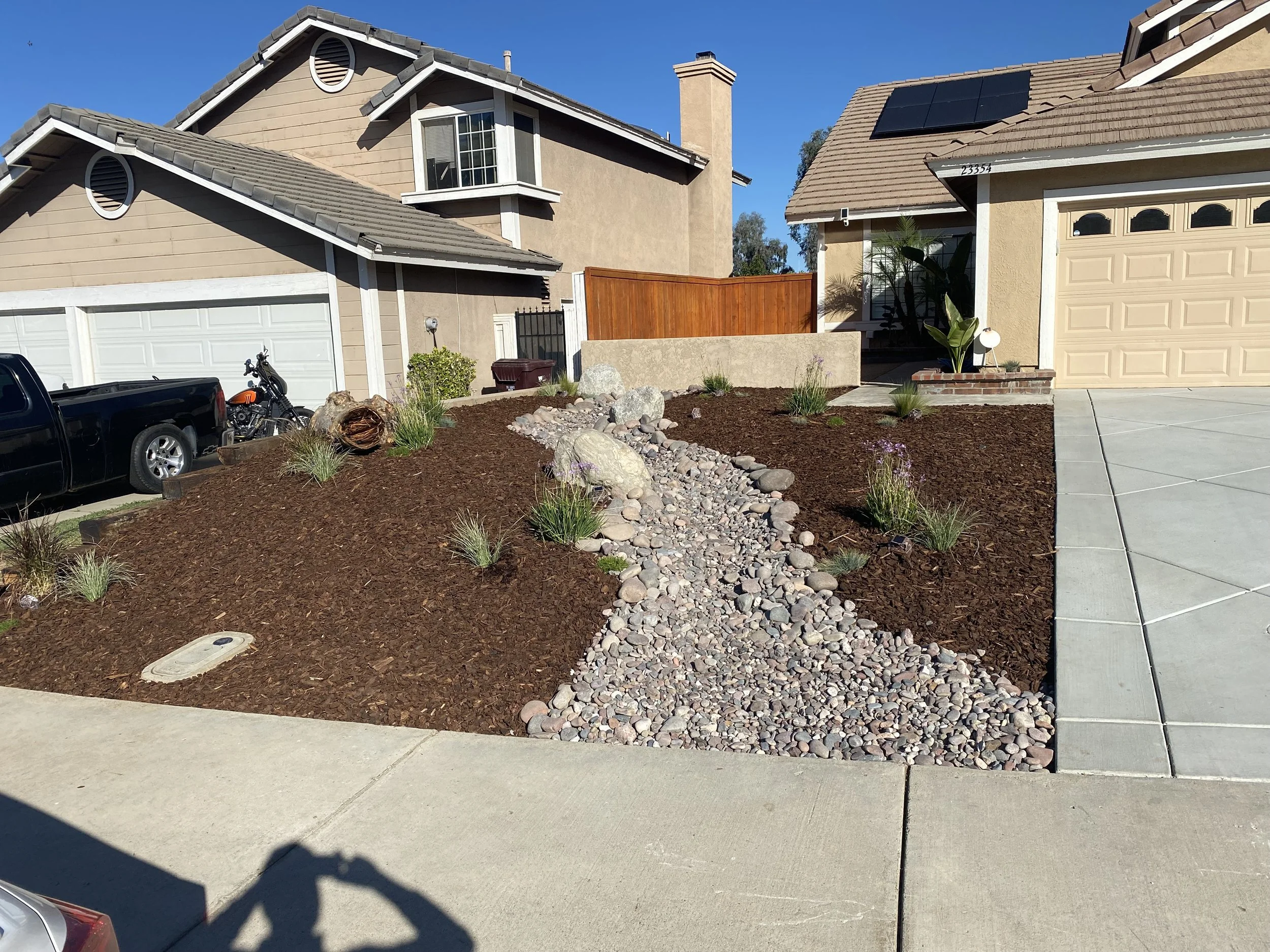 Front yard landscaping with a dry creek bed made of small rocks, mulch, small plants, and a large piece of driftwood between two houses with beige and brown exteriors, driveways, and a bright blue sky.