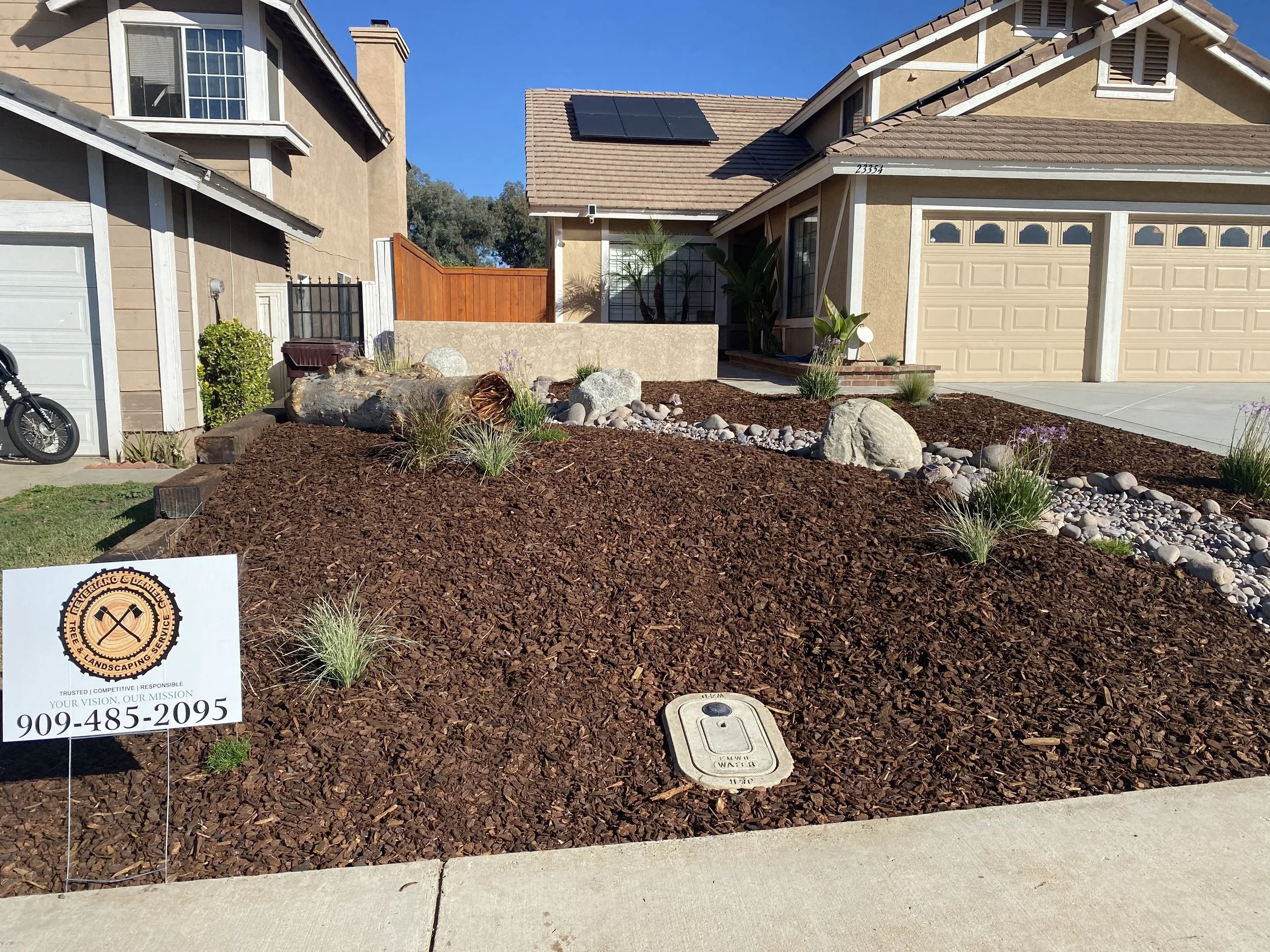 Front yard landscaping with mulch and small plants in front of a beige house with a garage, featuring rocks and large stones, and a sign for a landscaping service.