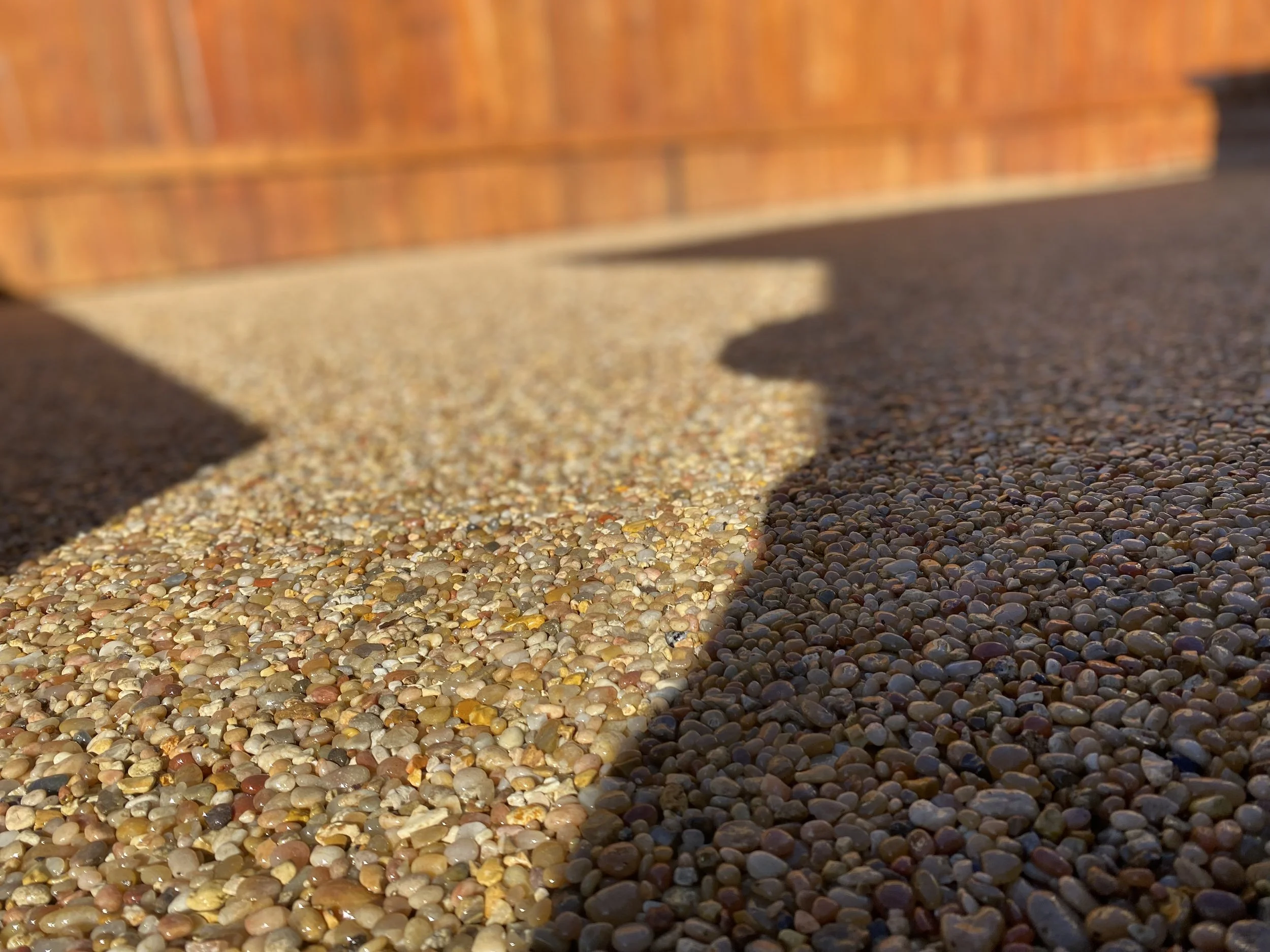 Close-up of a pebble-textured ground with shadows cast by nearby objects and a wooden fence in the background.