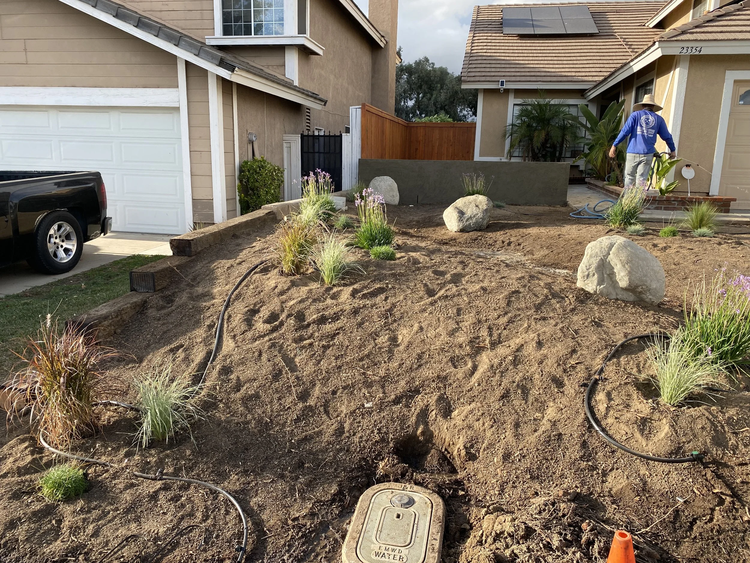Residential front yard landscaping in progress, featuring large decorative rocks, planted grasses, and a person working on the steps of a house.