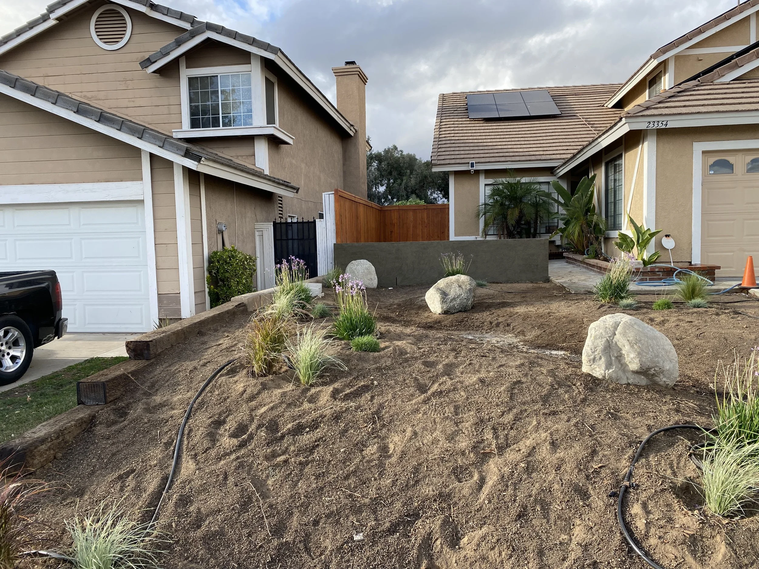 Front yard of a residential house under landscaping, with large rocks, plants, and exposed soil, with neighboring houses on each side and a cloudy sky.