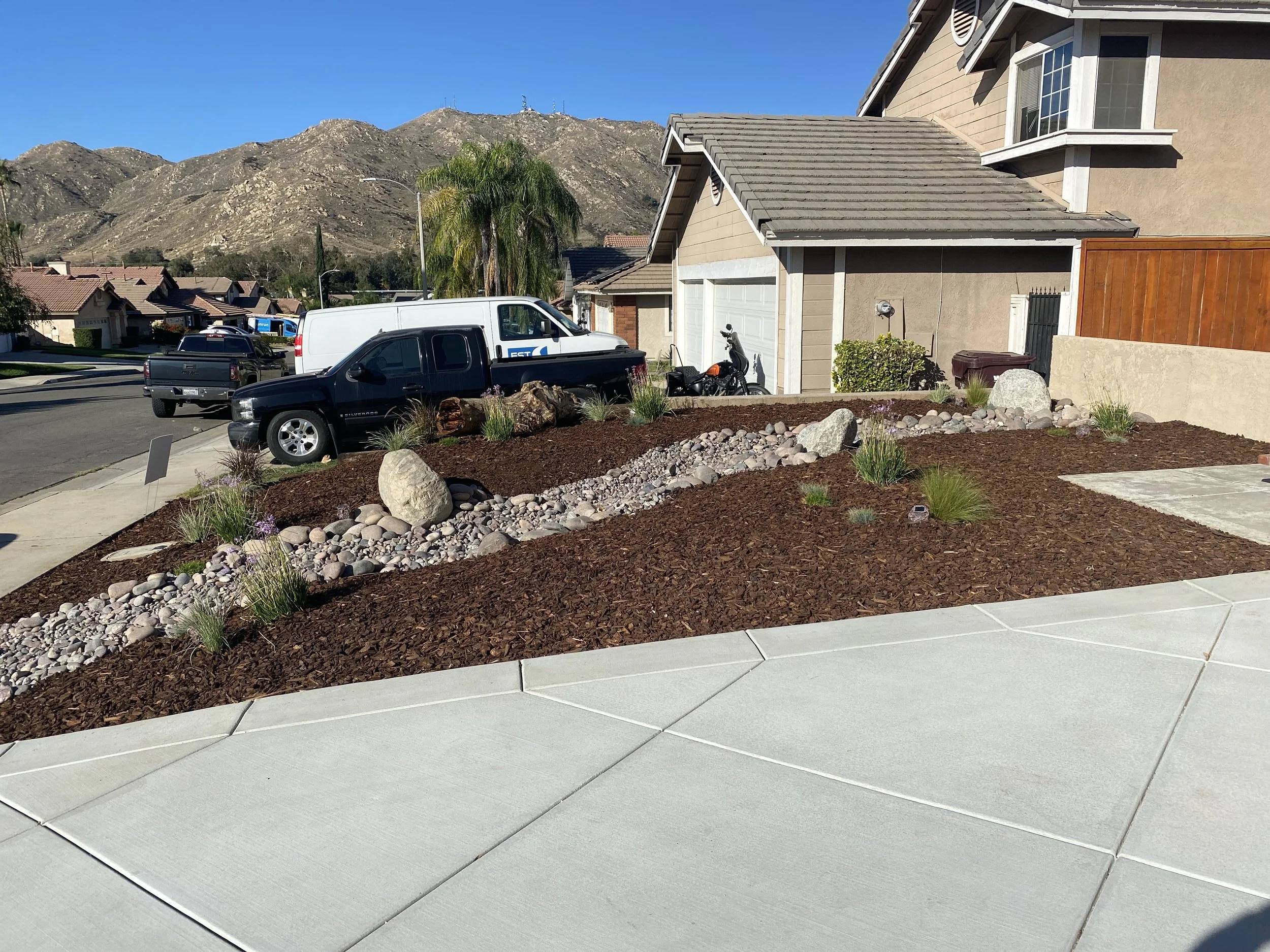 Residential front yard with landscaped rocks, mulch, and drought-tolerant plants, with houses and mountains in the background.