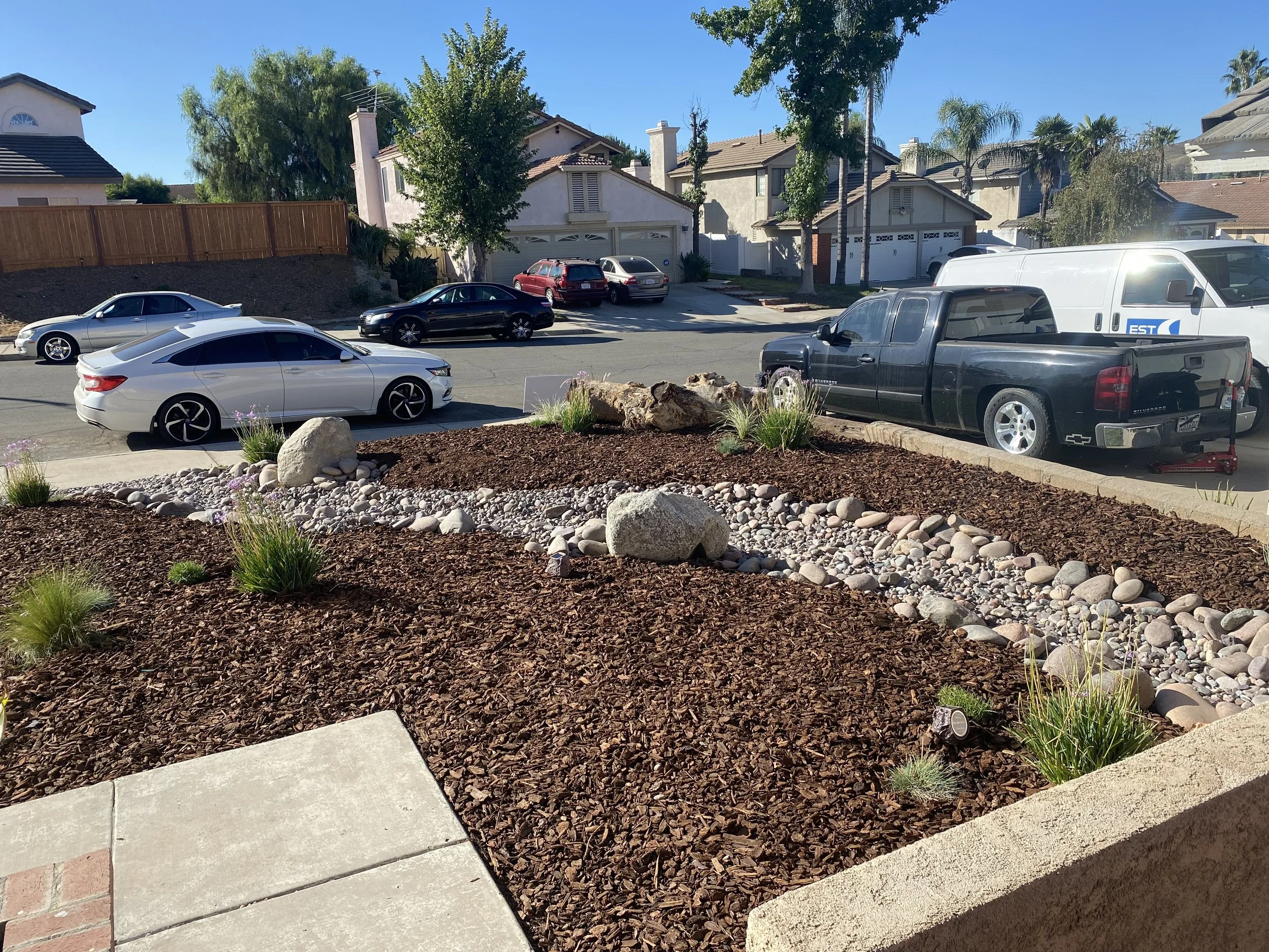 Residential street view showing parked cars, houses, trees, and a landscaped front yard with rocks and mulch.