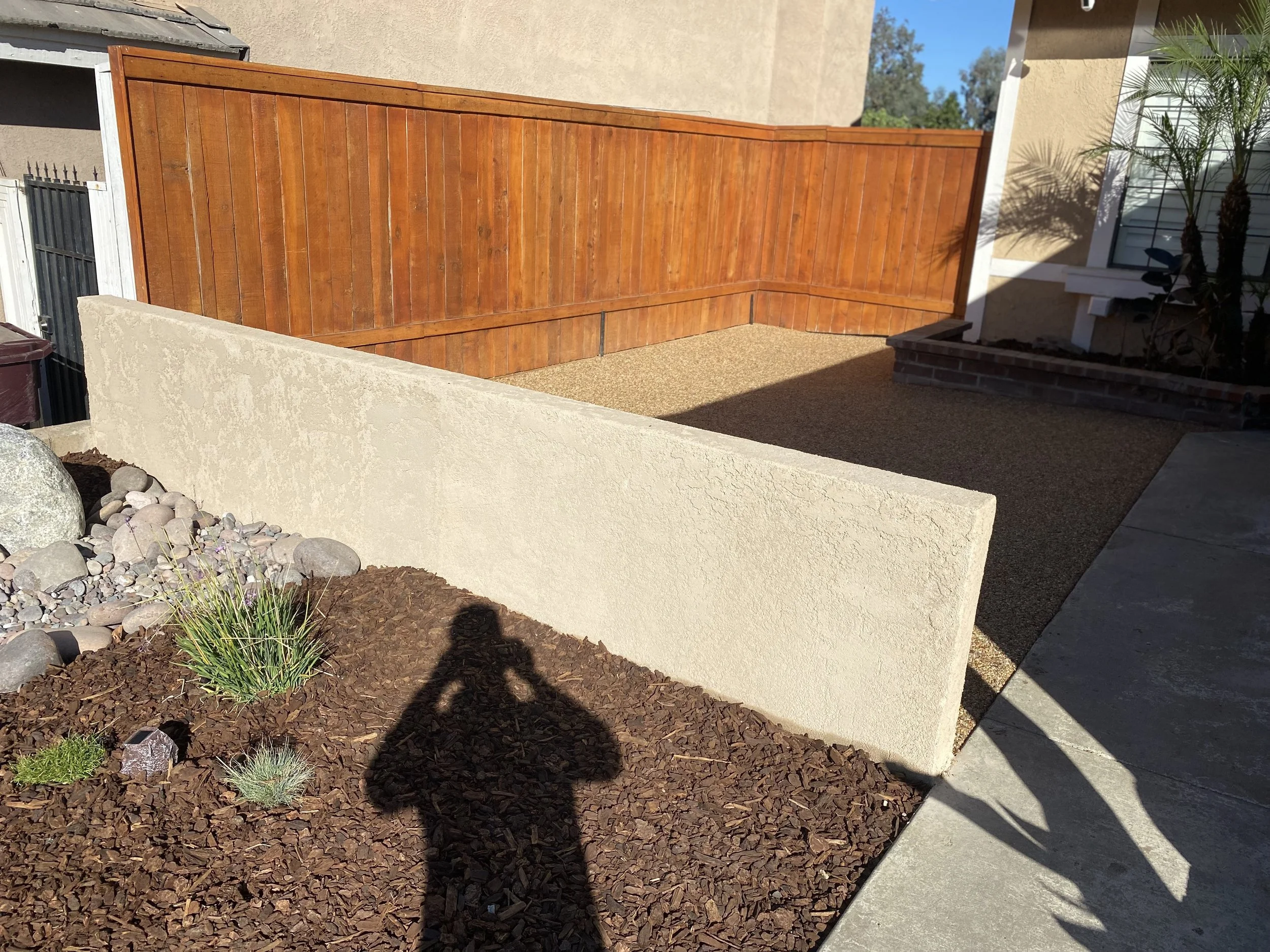 A backyard area with a beige concrete wall, a wooden privacy fence, gravel ground, and some plants, with shadows cast by nearby objects and a person taking the photo.
