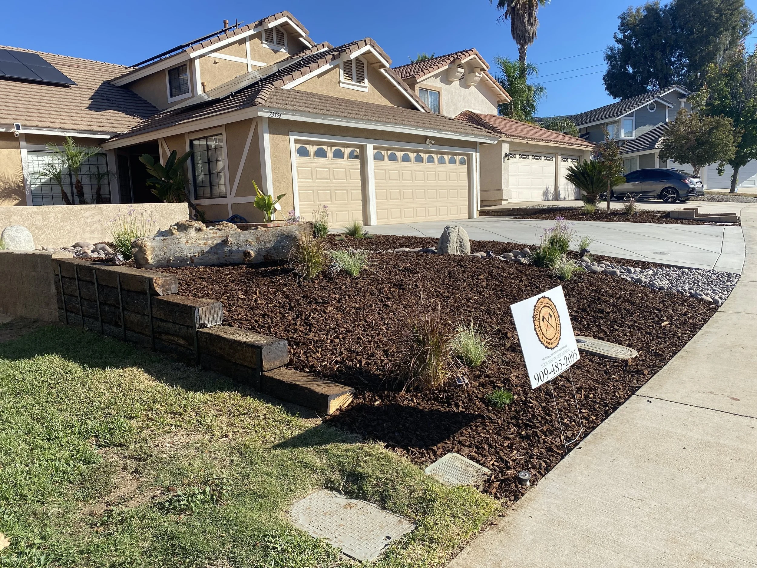 Front yard of a house with freshly landscaped garden, mulch, rocks, plants, a sign with a logo and phone number, and a driveway leading to a garage.
