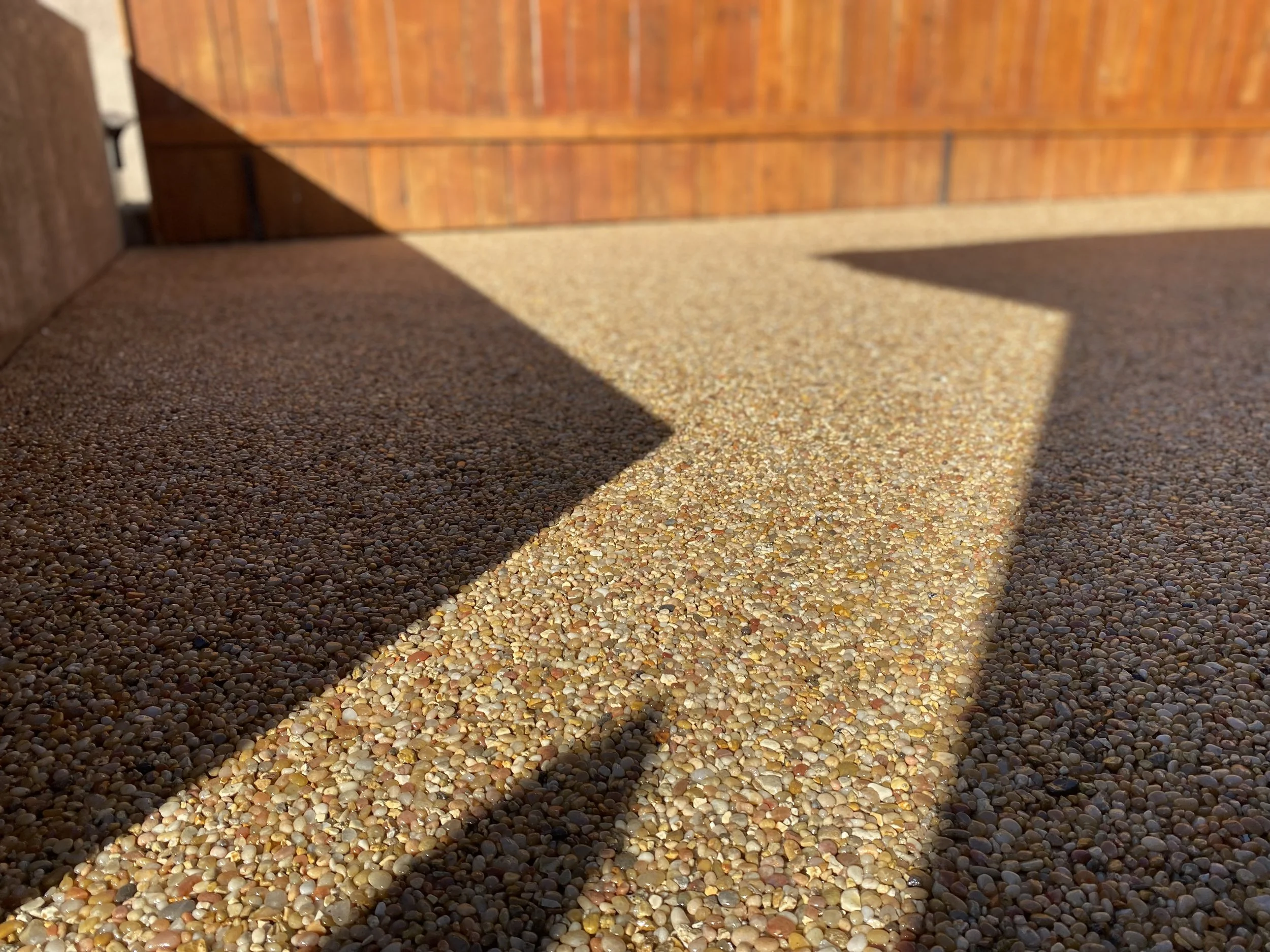 Close-up view of a pebble-textured driveway with shadows cast by a wooden fence or wall in the sunlight.