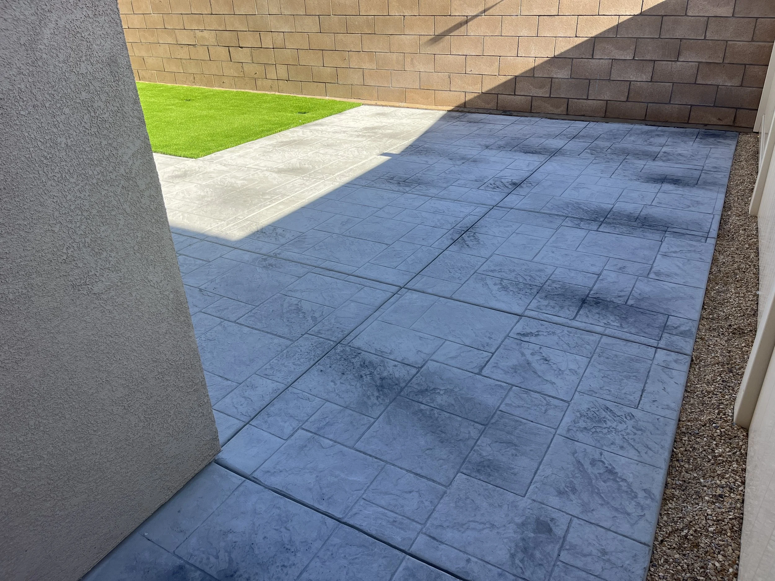 Photo of a small backyard patio with light gray stamped concrete pavers, a bit of green lawn, a brick wall, and a beige stucco wall with a shadow covering part of the patio.