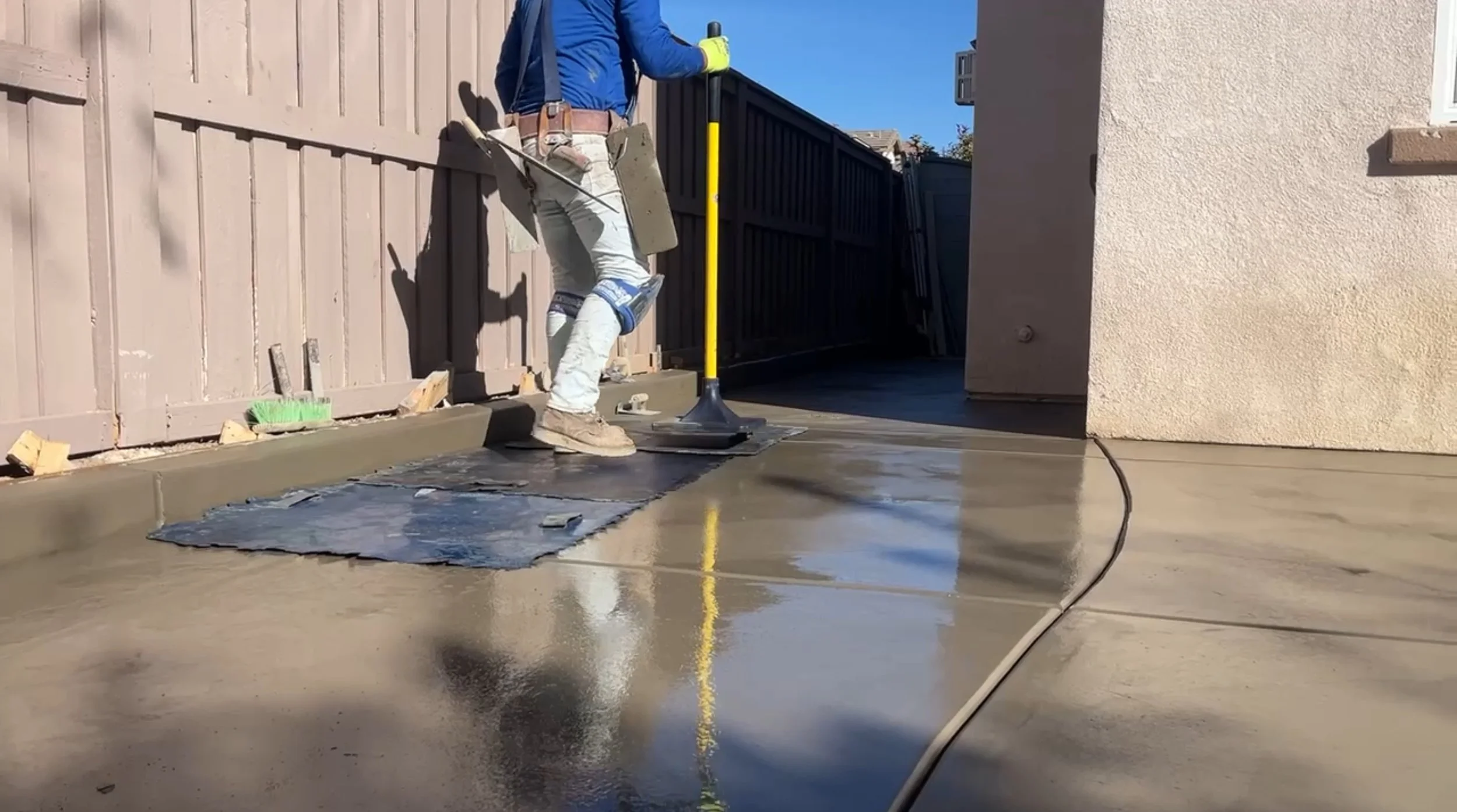 Worker is laying concrete on a sidewalk with a tamper and a yellow-handled tool, next to a wooden fence and house.