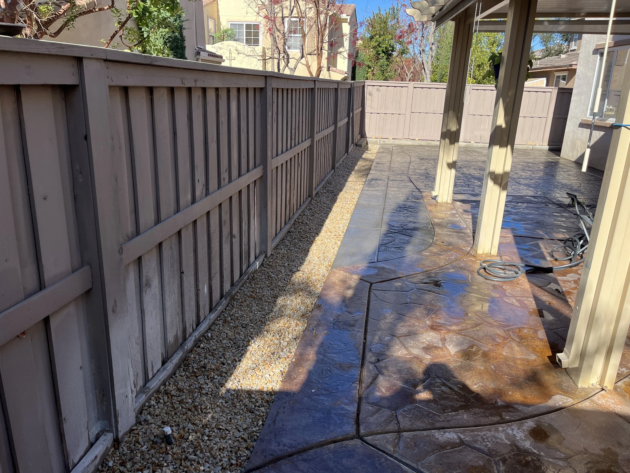 Backyard patio with a stamped concrete surface, a side gravel strip, and a wooden privacy fence. Part of a covered patio with support posts is visible on the right.