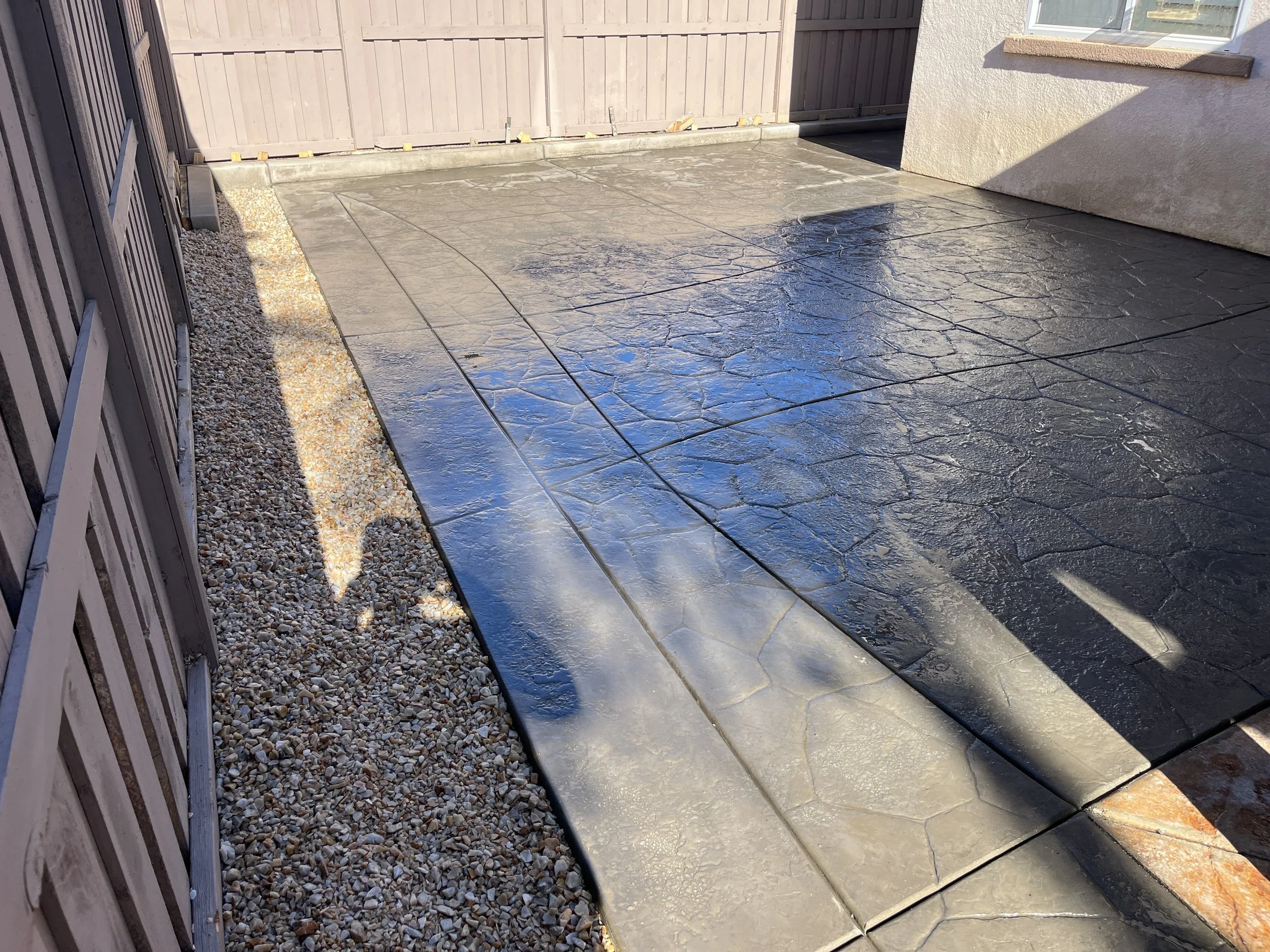 Freshly poured concrete patio with decorative cracks, bordered by gravel on one side and a wooden fence and house wall on the other.