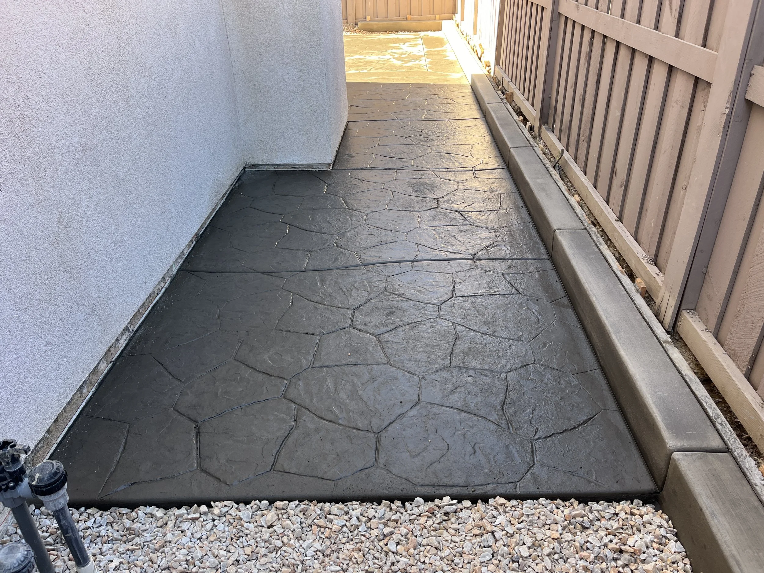 Concrete patio with stamped pattern, bordered by gravel and edged with metal and wooden fencing.