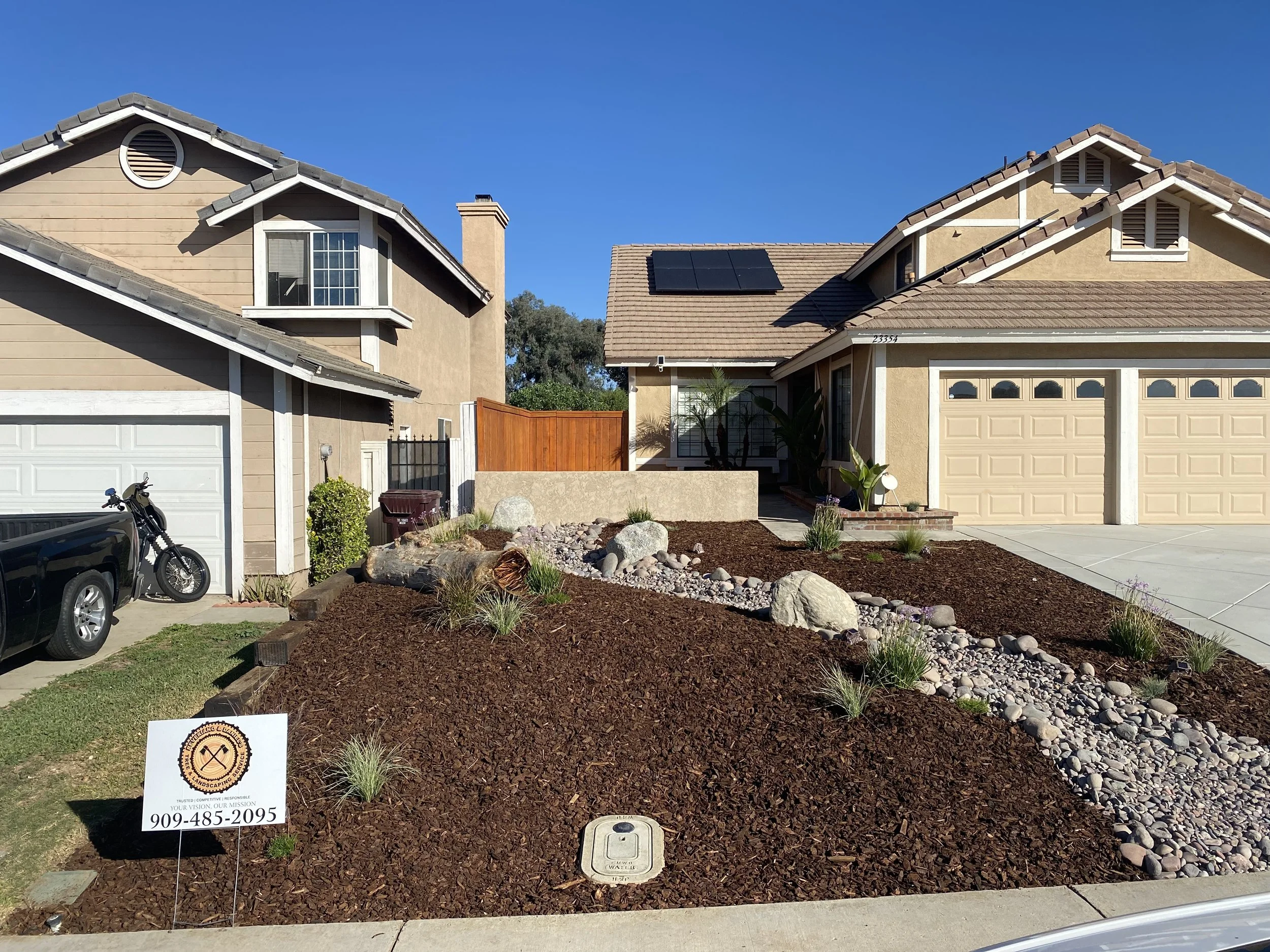 Front yard of a beige house with landscaping including rocks and mulch, a small sign with a logo and phone number, a black motorcycle, and neighboring houses with solar panels on the roof.