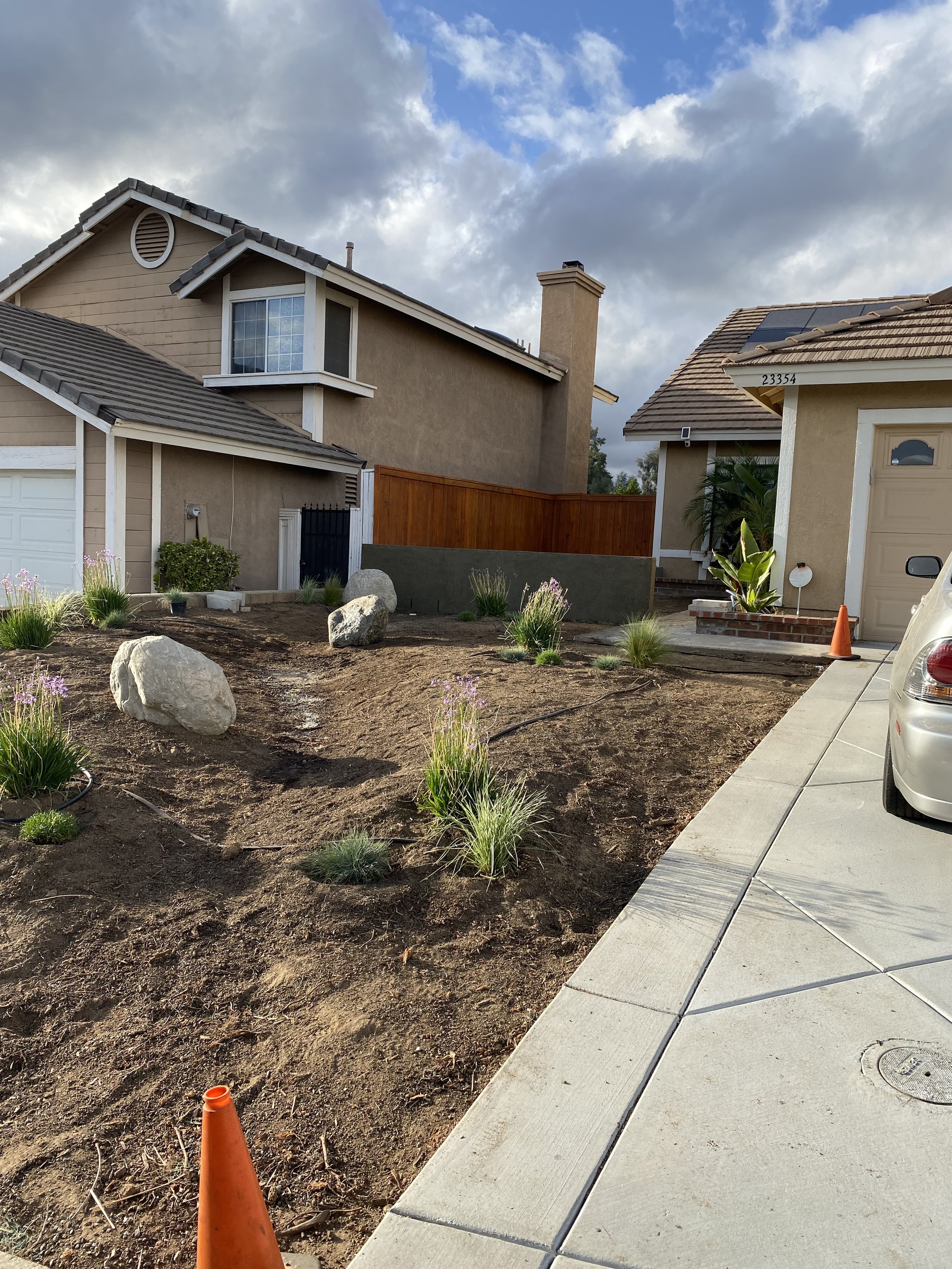 A residential front yard undergoing landscaping, with bare soil, sprouting plants, decorative rocks, and orange construction cones, with neighboring houses and a cloudy sky in the background.