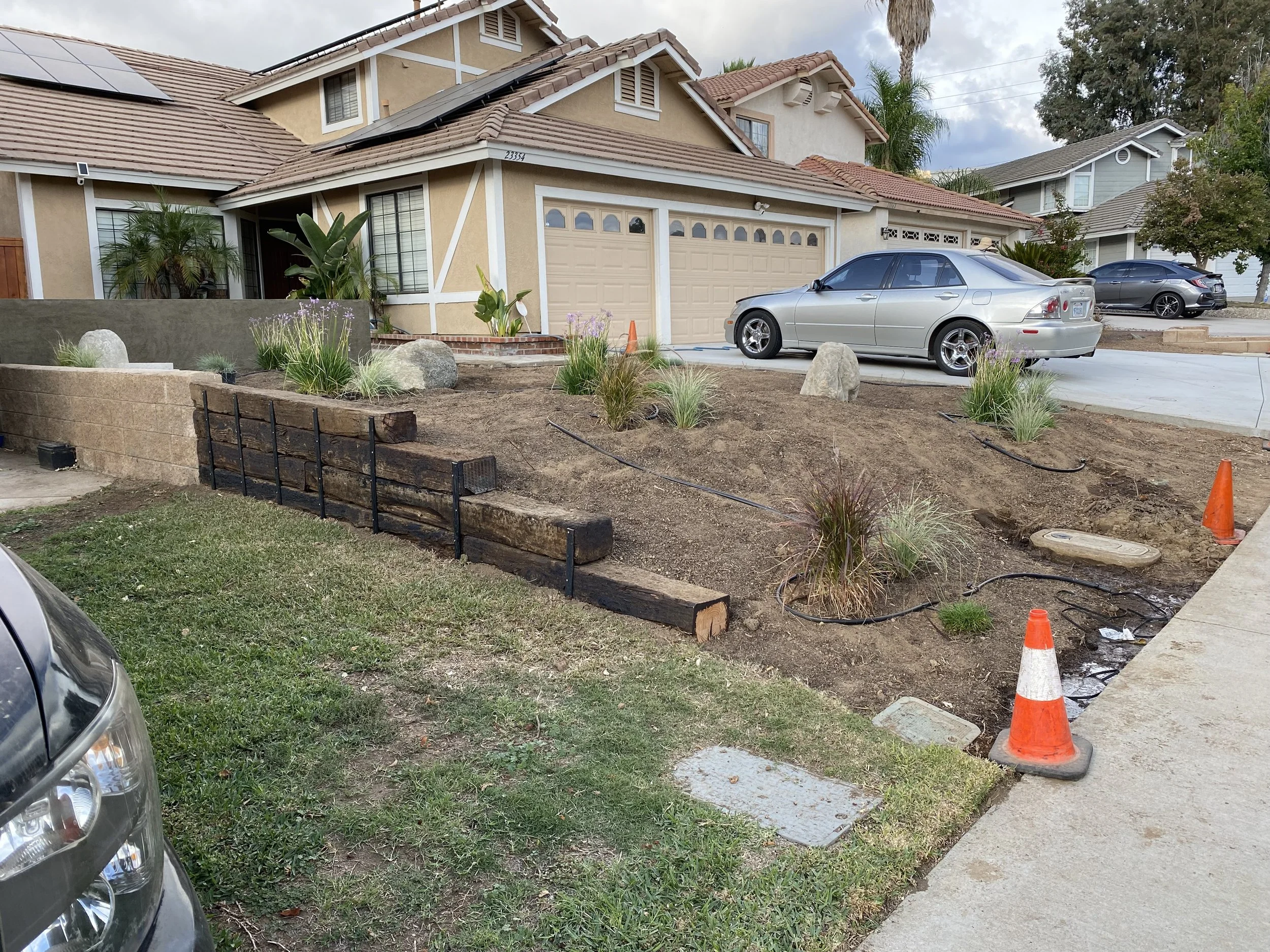 Front yard of a house under renovation with a layer of dirt and plants, orange construction cones, and exposed pipes.