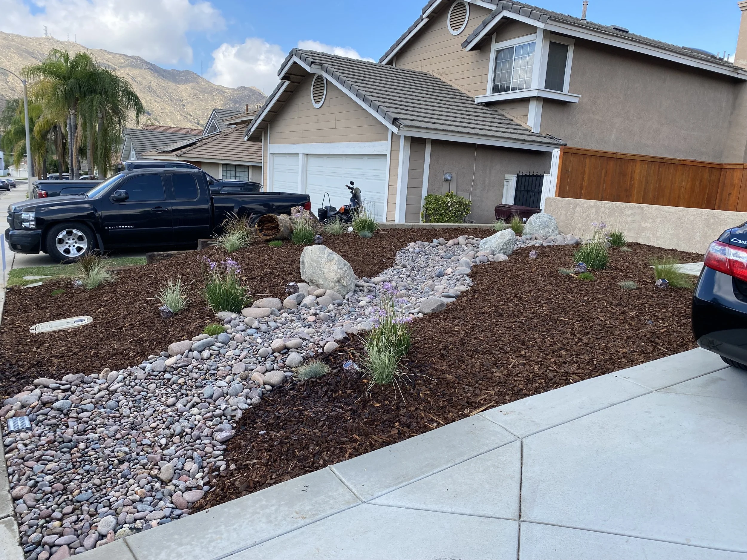 Residential front yard landscaping with mulch, rocks, and sparse plants. A concrete driveway broom Finish in Moreno Valley 