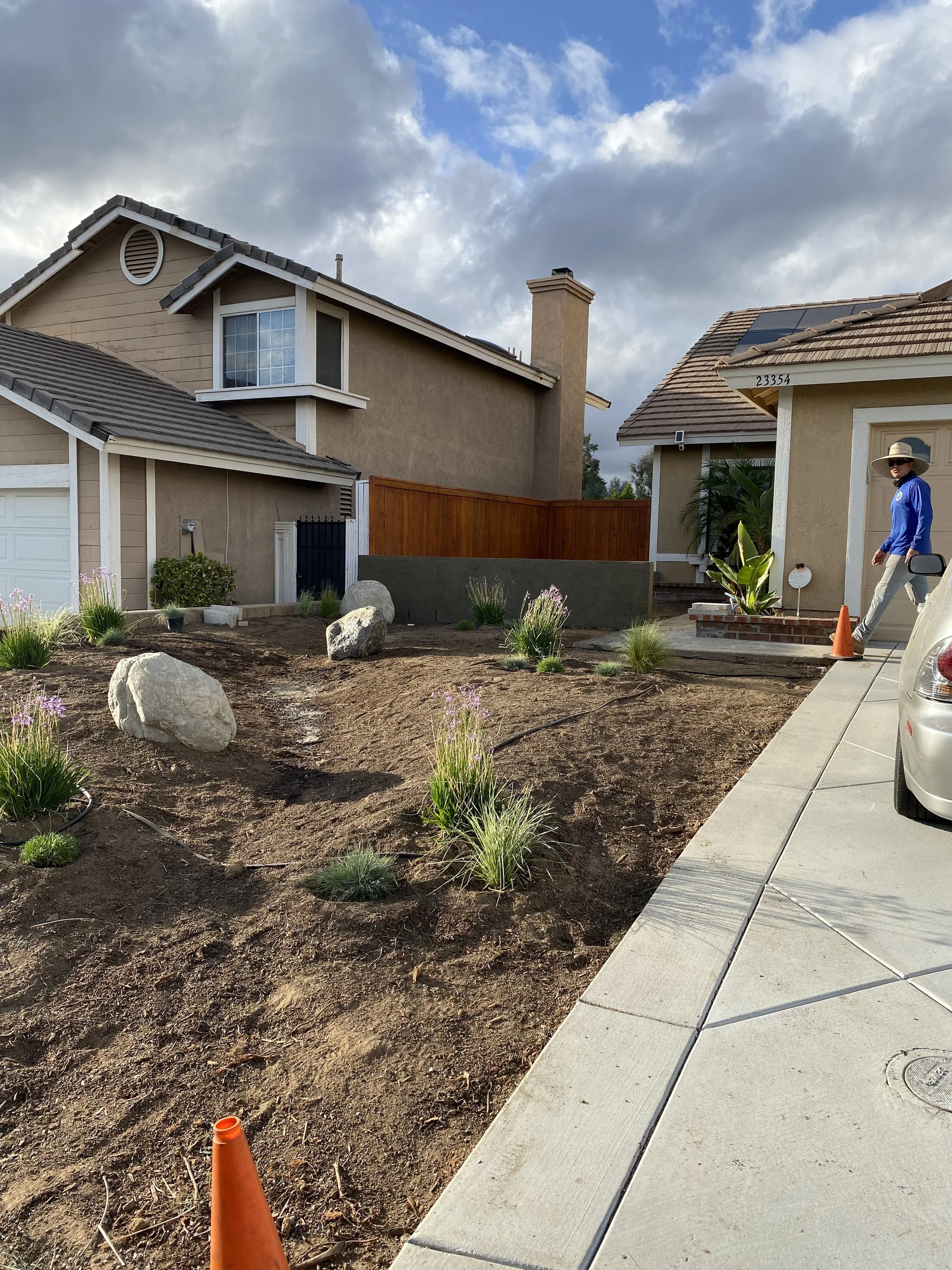 A newly landscaped front yard with small plants and decorative rocks, a concrete driveway, and two houses in the background. A person wearing a hat and sunglasses is walking near the house on the right, and an orange traffic cone is on the sidewalk.