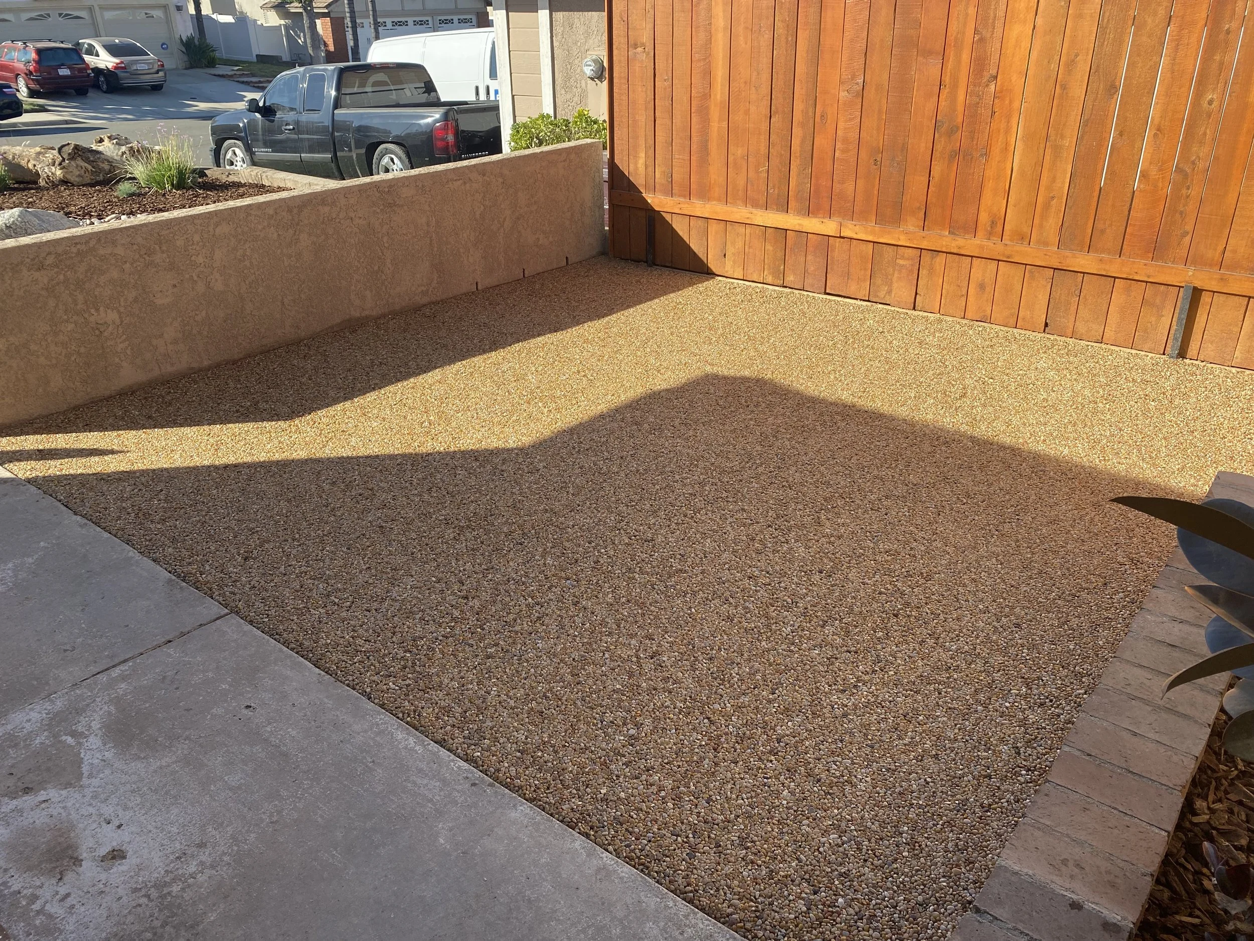 Small yard with pebble gravel surface, enclosed by a wooden fence and a low concrete wall, with parked cars visible beyond.