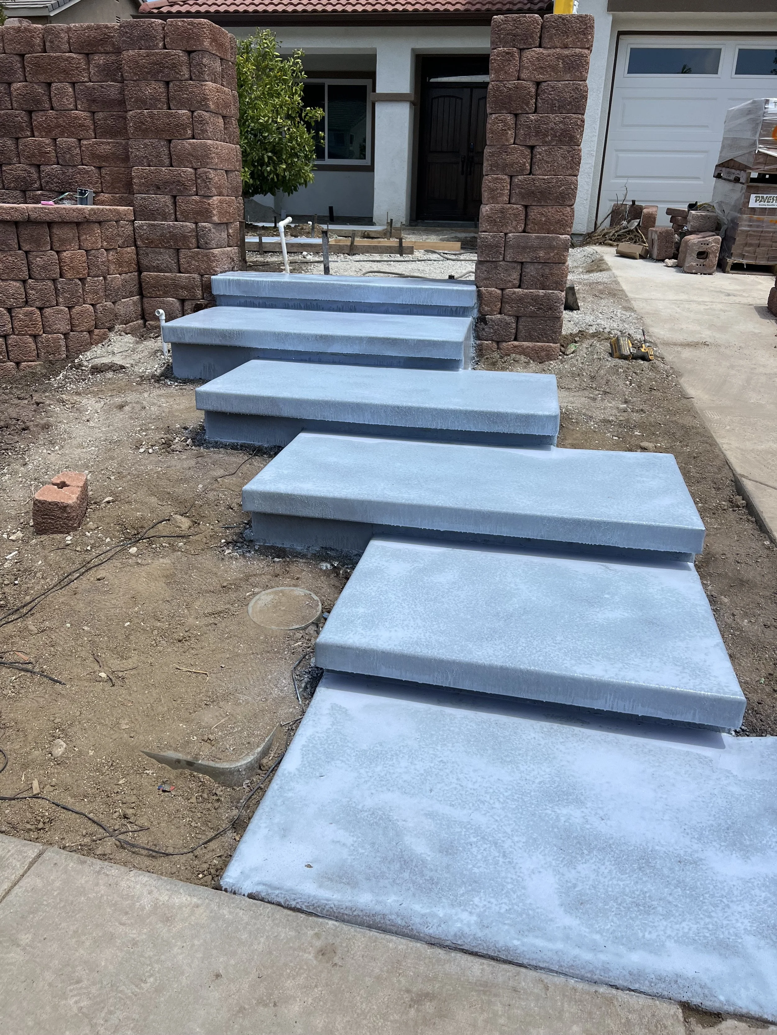 Concrete steps under construction leading to the front door of a house, with a brick wall on either side and construction materials nearby.