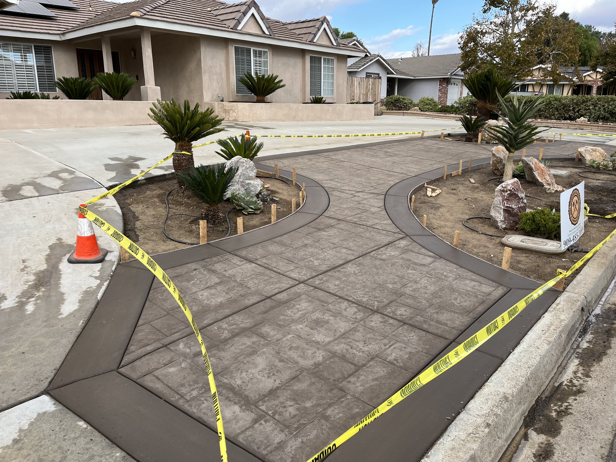 Landscaping in progress outside a residential house with newly installed curved concrete pathways, decorative rocks, and plants, surrounded by caution tape.