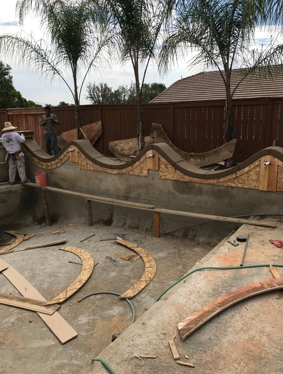 Construction of a wave-shaped concrete wall with wooden framing and decorative elements, with workers and palm trees in the background.