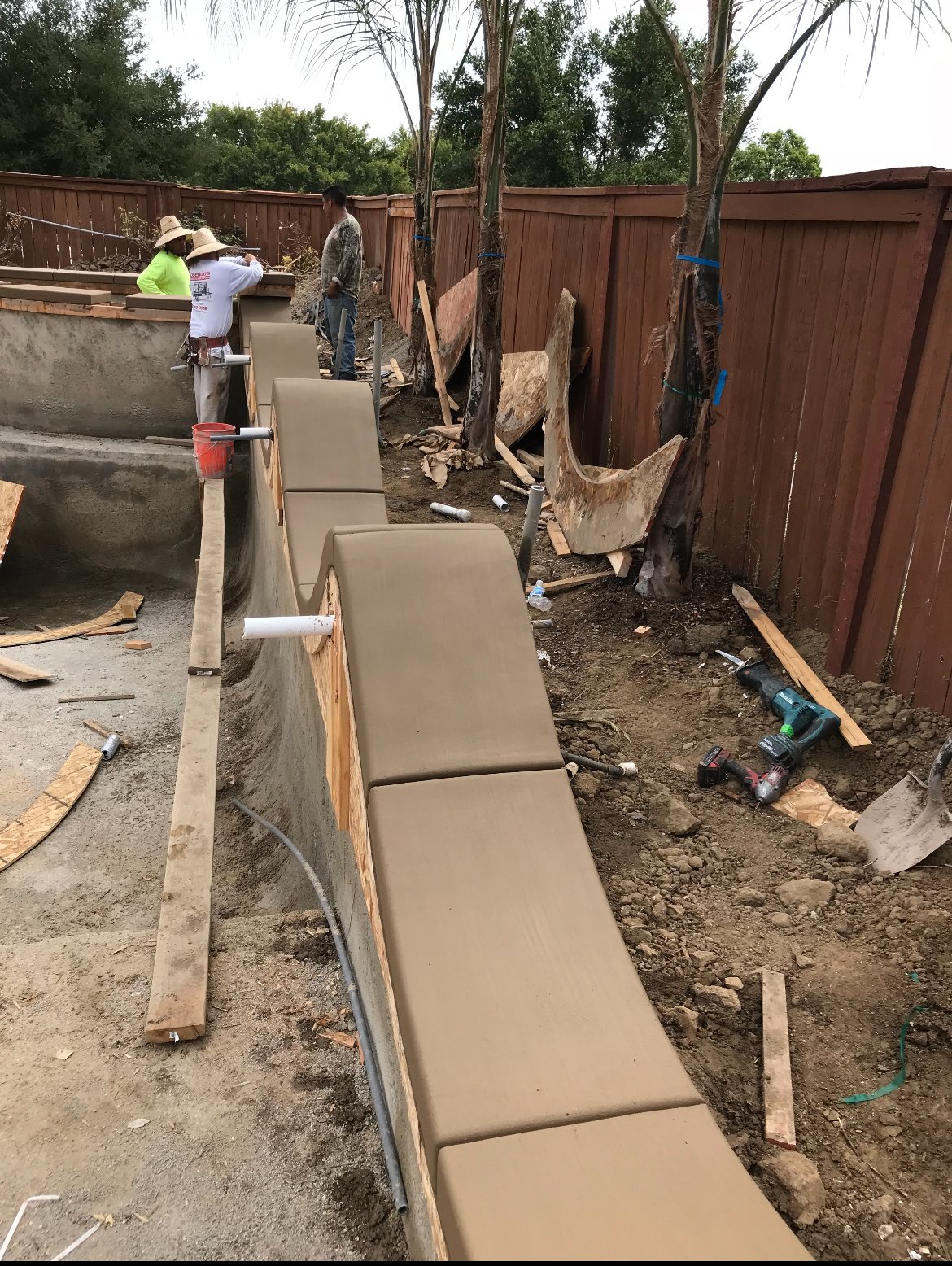 Construction workers building a backyard water feature with a concrete waterfall and tiered benches, surrounded by wooden fencing and palm trees.