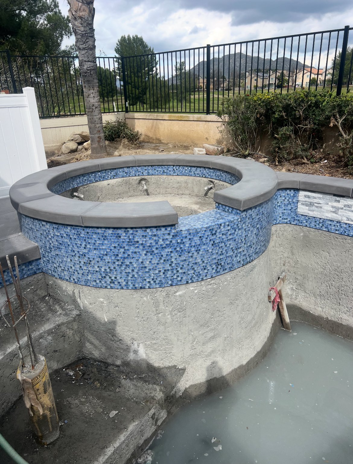 Under construction hot tub with blue mosaic tiles and gray stone ledge, surrounded by dirt and construction materials, fenced backyard with trees and mountains in the background.