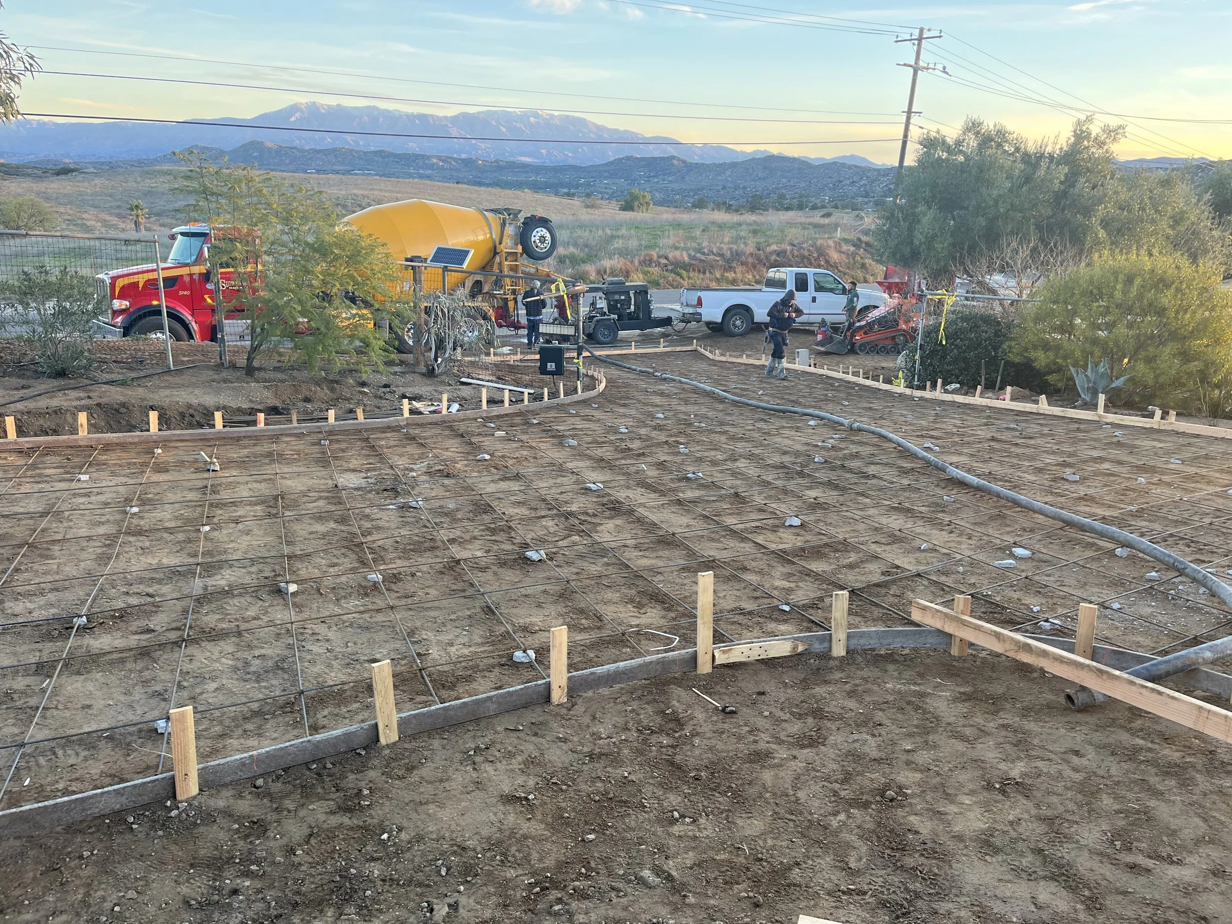 Construction site for a new building with rebar grid in the foreground, construction workers, a concrete mixer truck, and utility trucks in the background, with a scenic mountainous landscape in the distance.