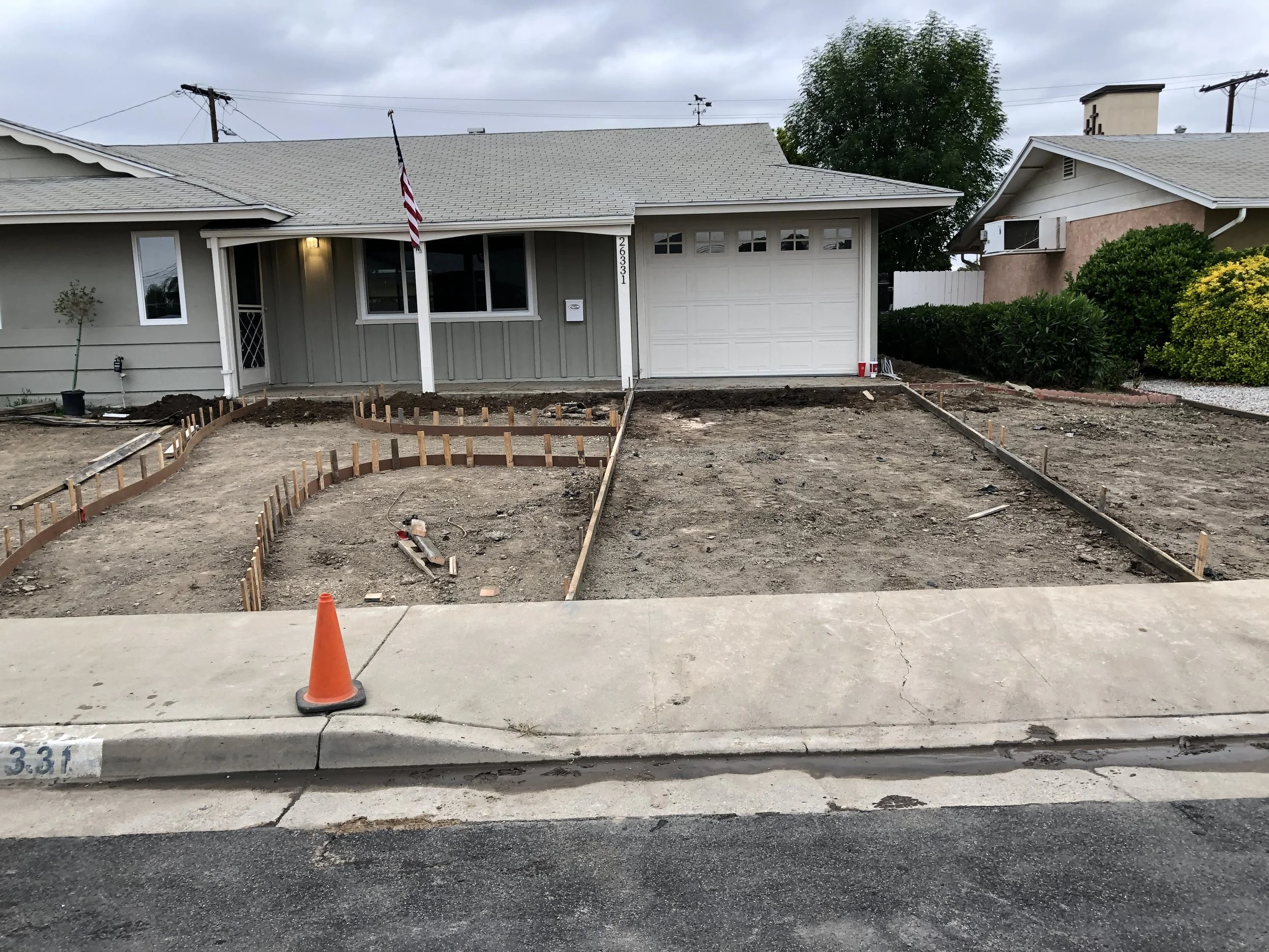 Front yard of a house under renovation, with dirt and wooden border frames for new landscaping, a cone on the sidewalk, and a house with a light gray exterior and attached garage.