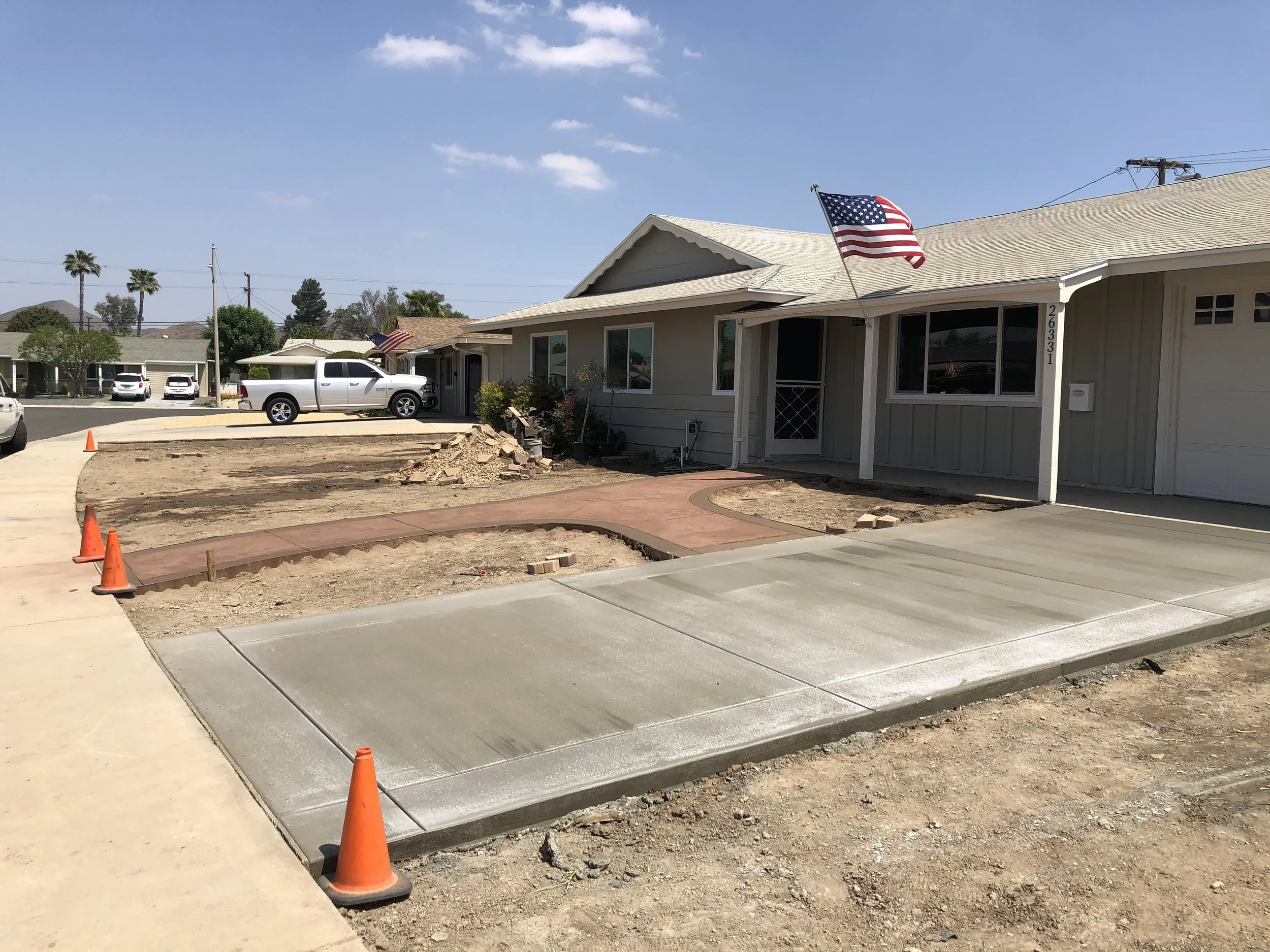 Construction of a new sidewalk in front of a house with an American flag, with traffic cones marking the area, and parked cars in the background.
