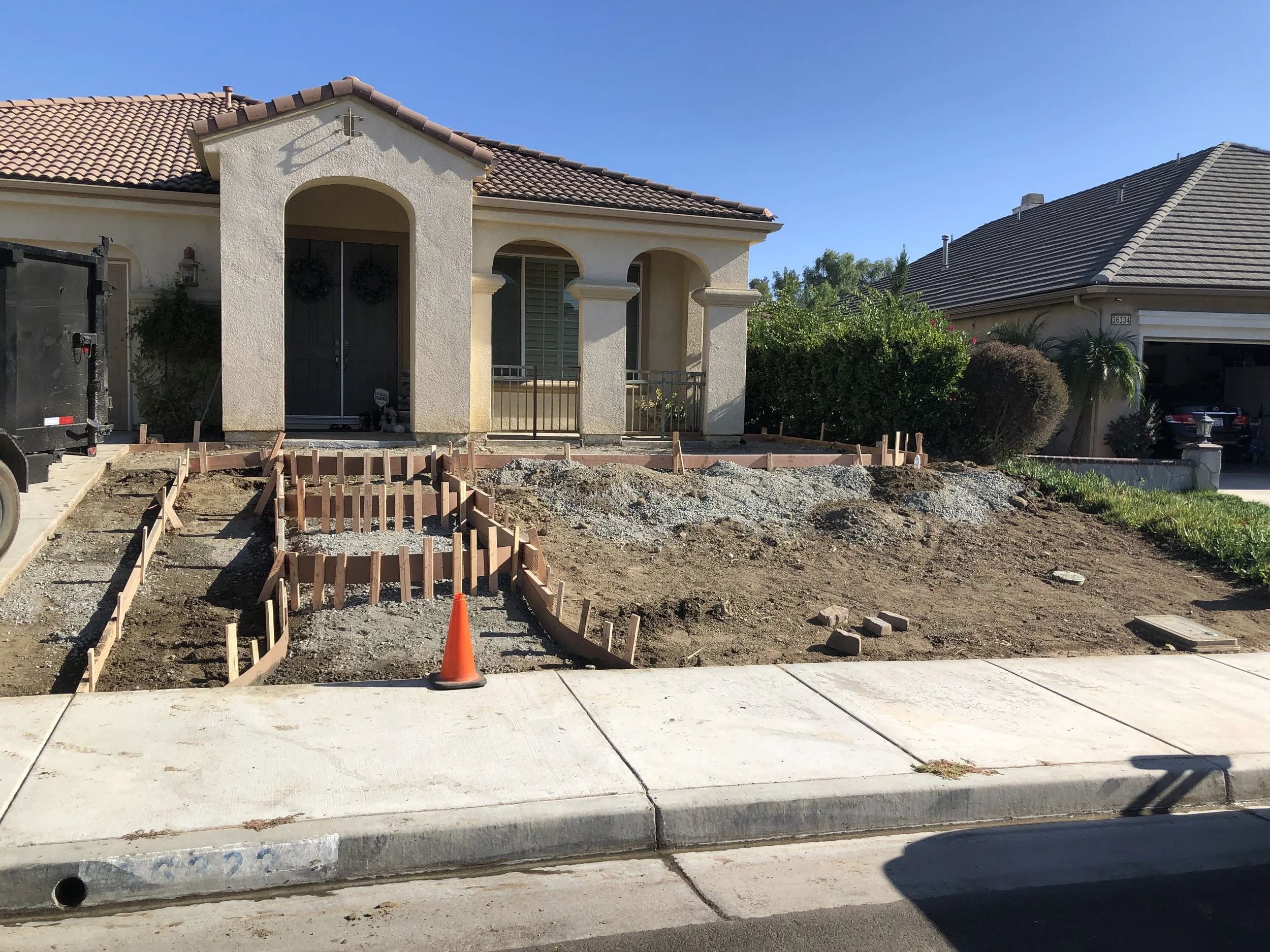 Front yard of a house under construction, with soil and wooden frames for a new walkway or garden bed, and a construction cone.