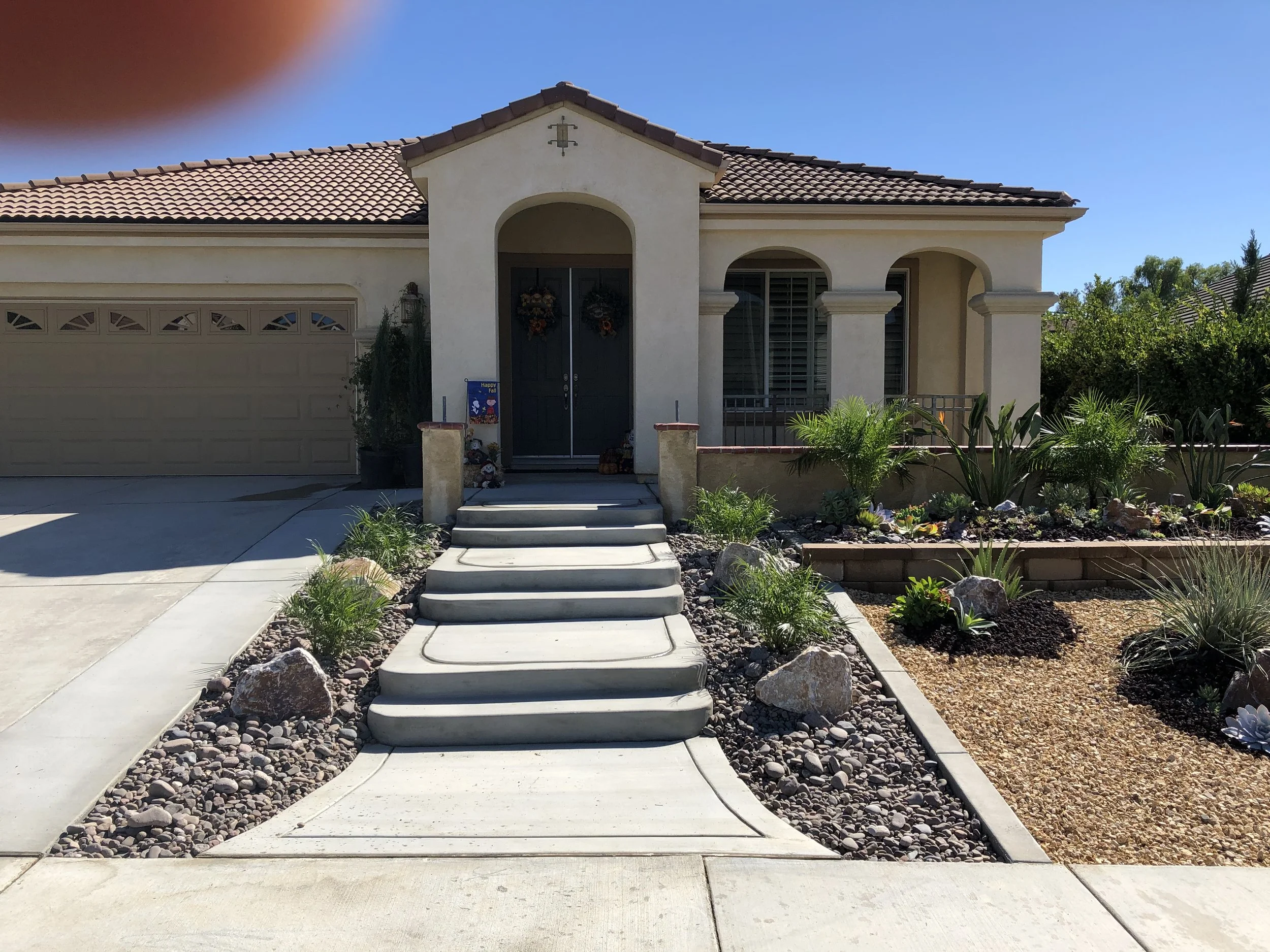 Front view of a house with a beige stucco exterior, front steps leading to dark double doors with wreaths, and a landscaped yard with rocks and green plants.