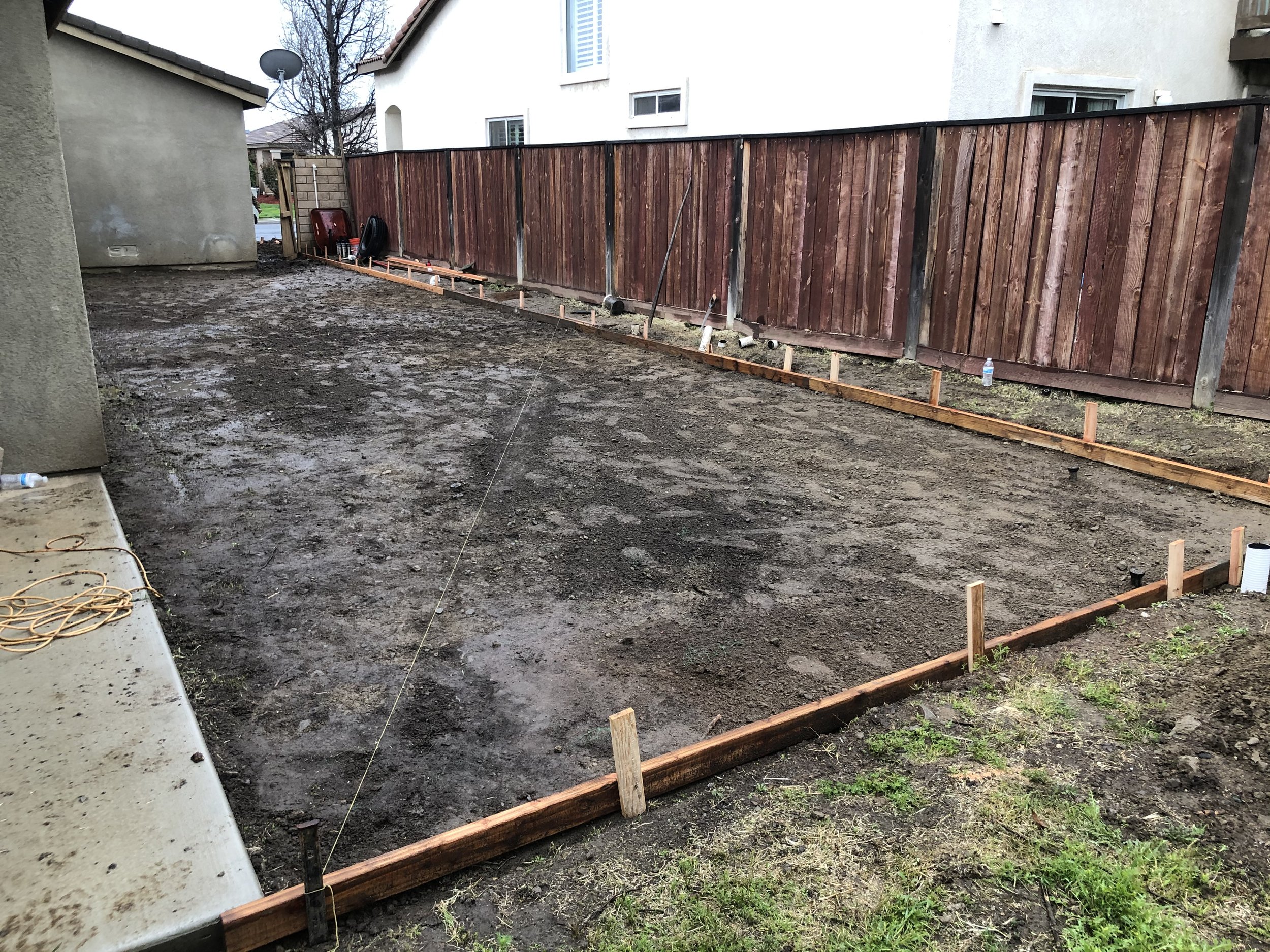 A backyard with a newly leveled ground, wooden border framing the area, and a wooden fence along the back. Construction tools and materials are present, with a plastic water bottle seen on the ground near the fence.