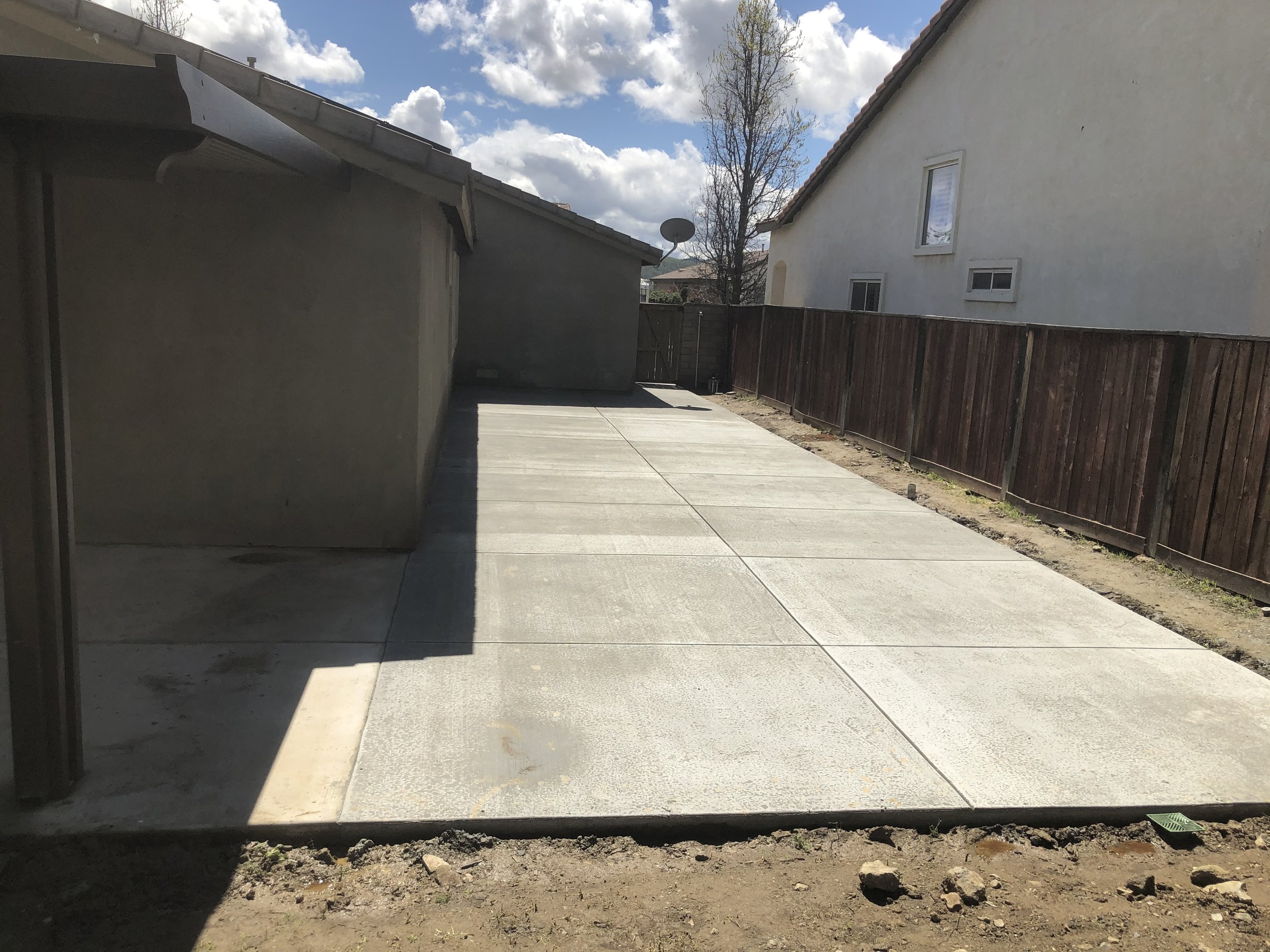 Freshly poured concrete patio in a backyard, with a wooden fence on one side and a house with a window and a satellite dish in the background.