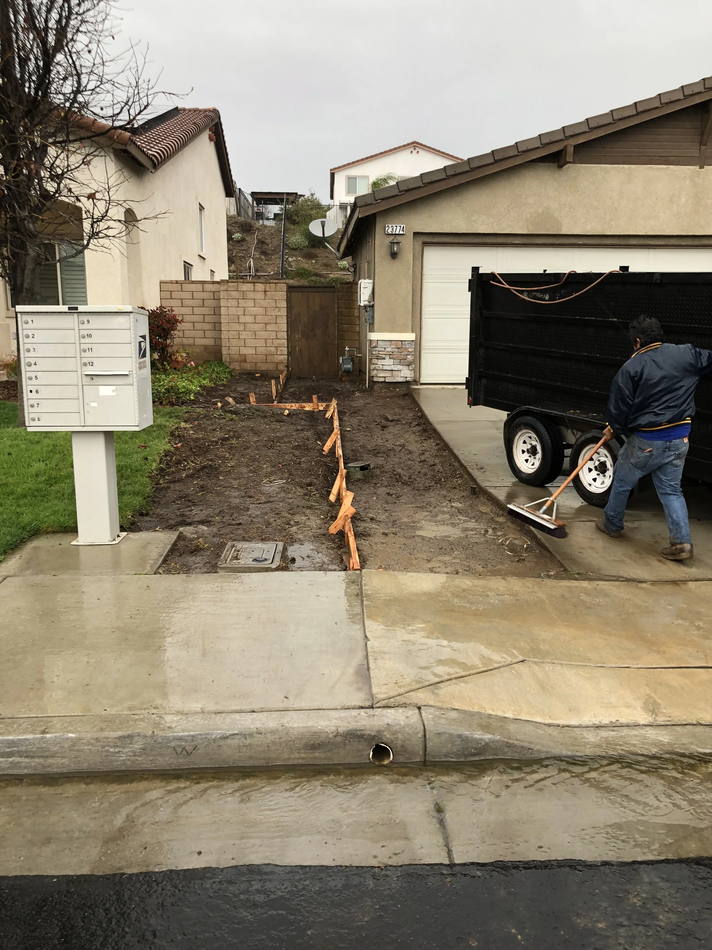 A person working on a front yard renovation, using a broom on a concrete driveway next to a black trailer, with a new garden bed area outlined by wooden supports in front of a beige house with a white garage door.