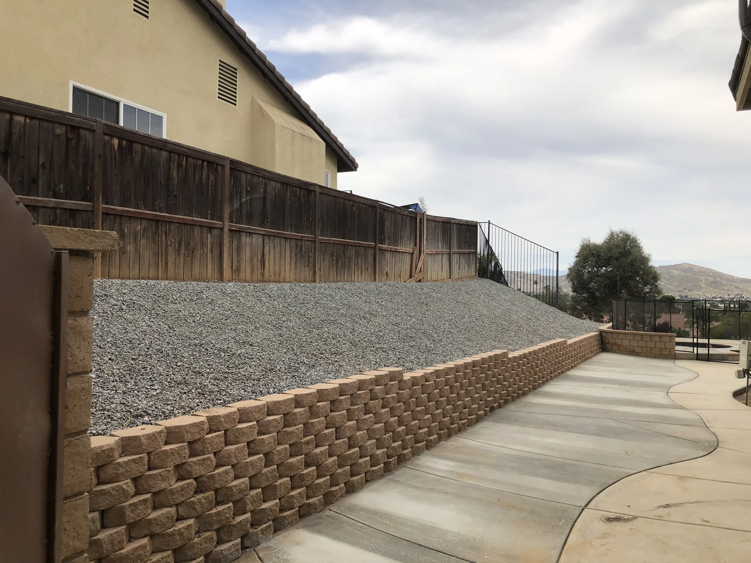 A backyard with a curved concrete patio, a retaining wall made of interlocking bricks, and a section with gravel and wood fencing. There is a hill and mountains in the background, with trees and a cloudy sky.