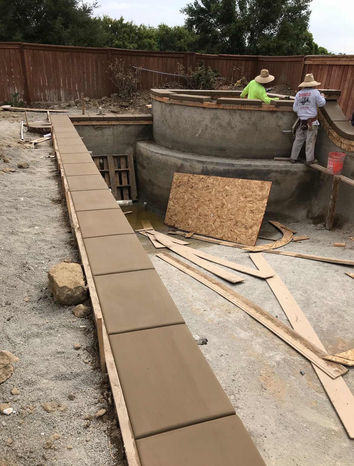 Construction workers building a swimming pool in a backyard, with tiled edges and a curved concrete wall, surrounded by a wooden fence.