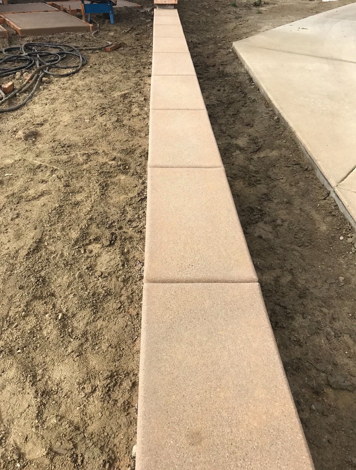 A newly installed sidewalk made of beige concrete slabs with joints, running alongside a dirt area and a larger concrete area on the right. Construction materials and tools are visible in the background.