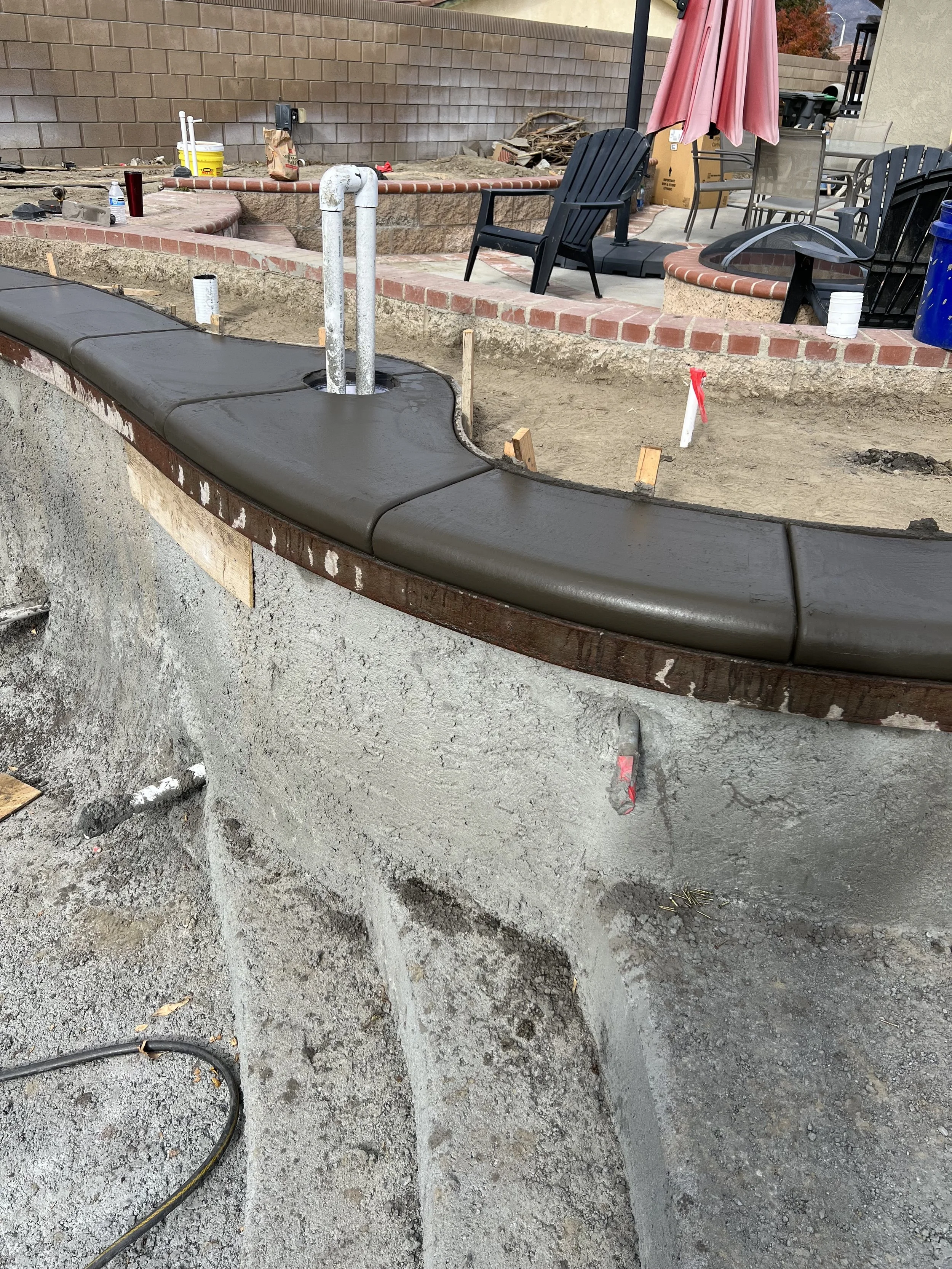 Under construction of a pool or hot tub with a concrete edge, newly poured coping, and plumbing installed, with patio furniture and a pink umbrella in the background.