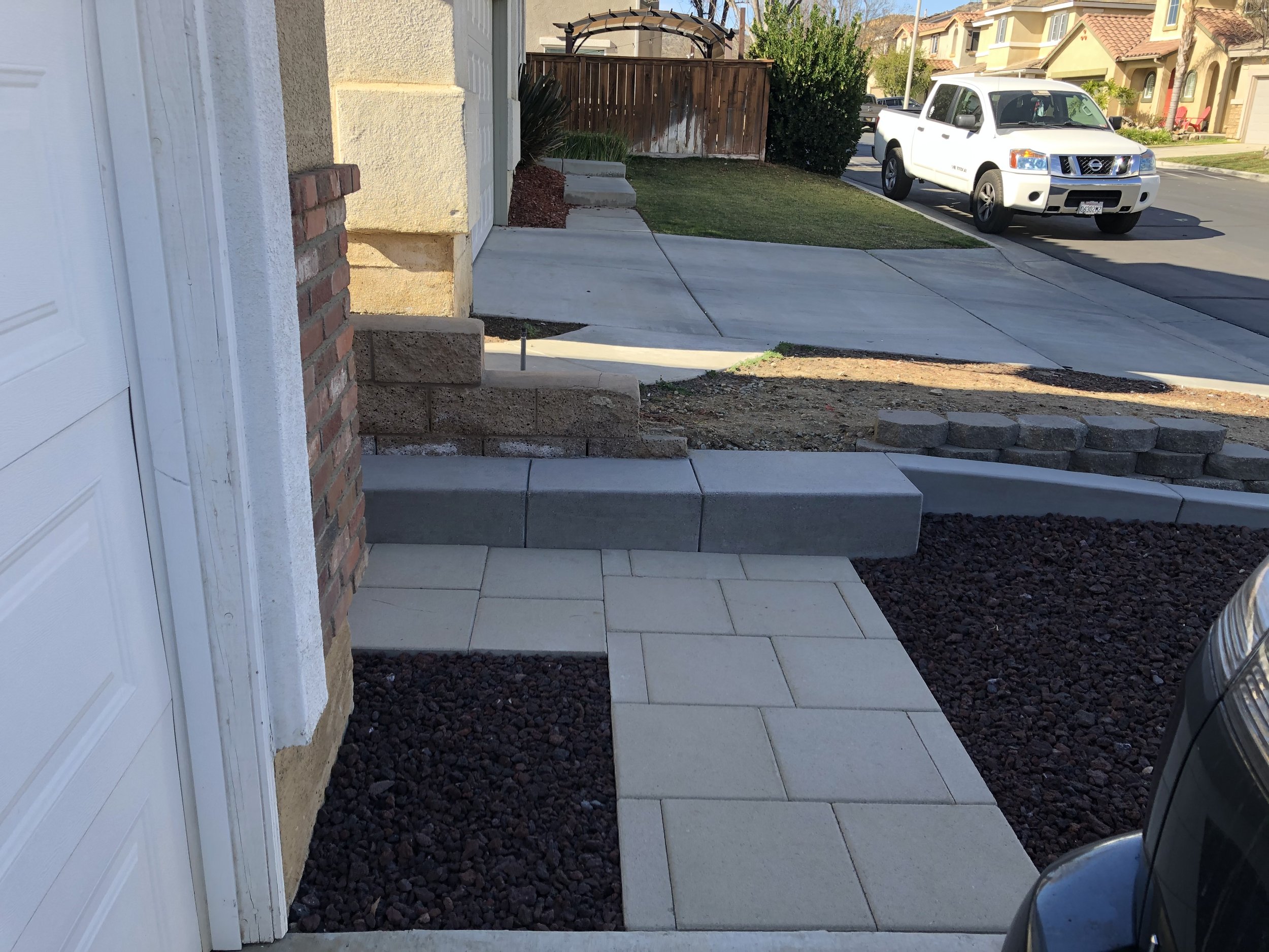 View of a residential front yard under construction with a concrete walkway, brick and stone steps, and a pile of pavers, near the street with a white pickup truck parked in the driveway.