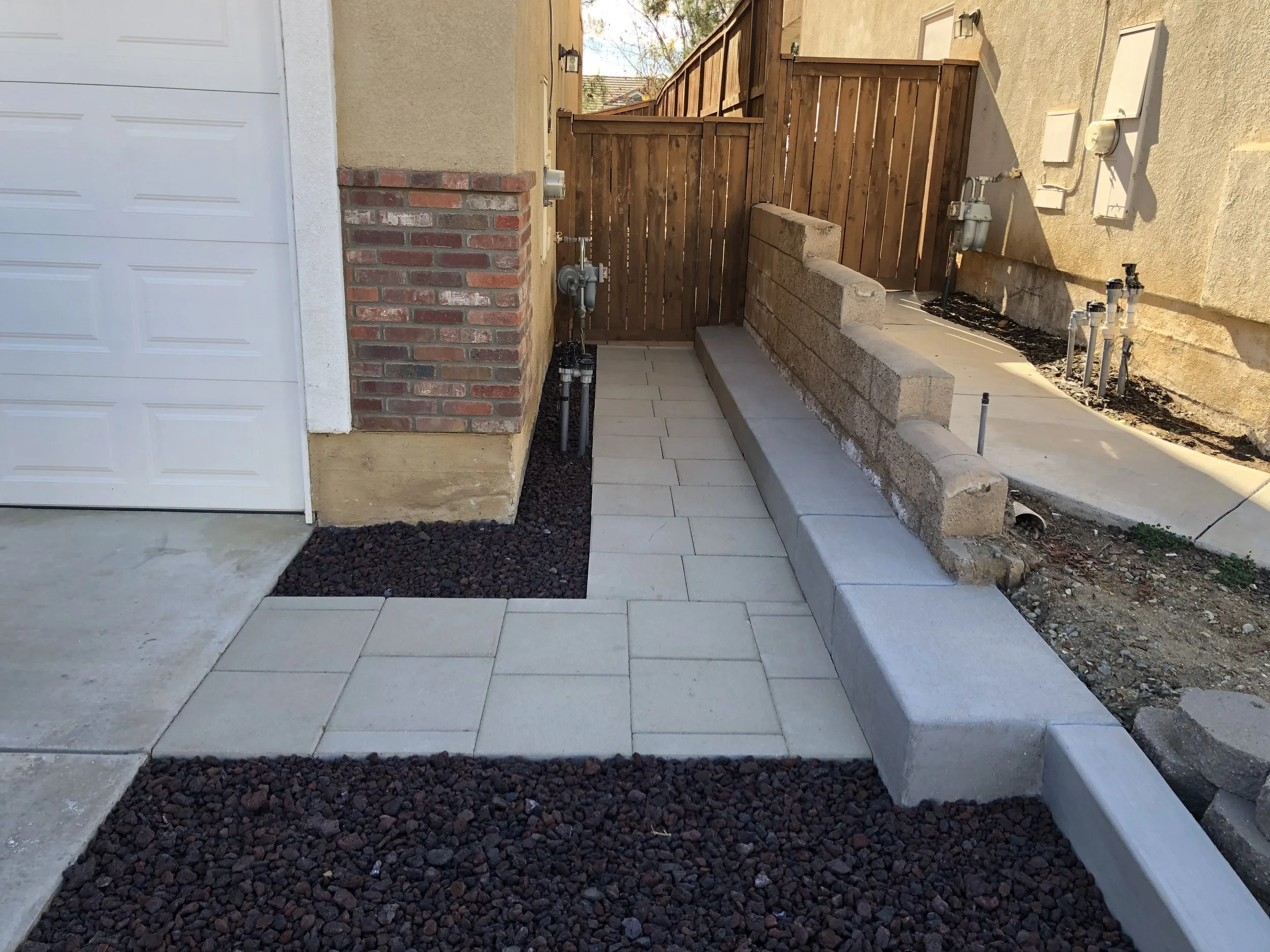 A newly paved backyard walkway with concrete pavers, bordered by black lava rocks, leading to a wooden gate in a fenced backyard. The alleyway features a brick wall, utility meters, and a small retaining wall made of concrete blocks.